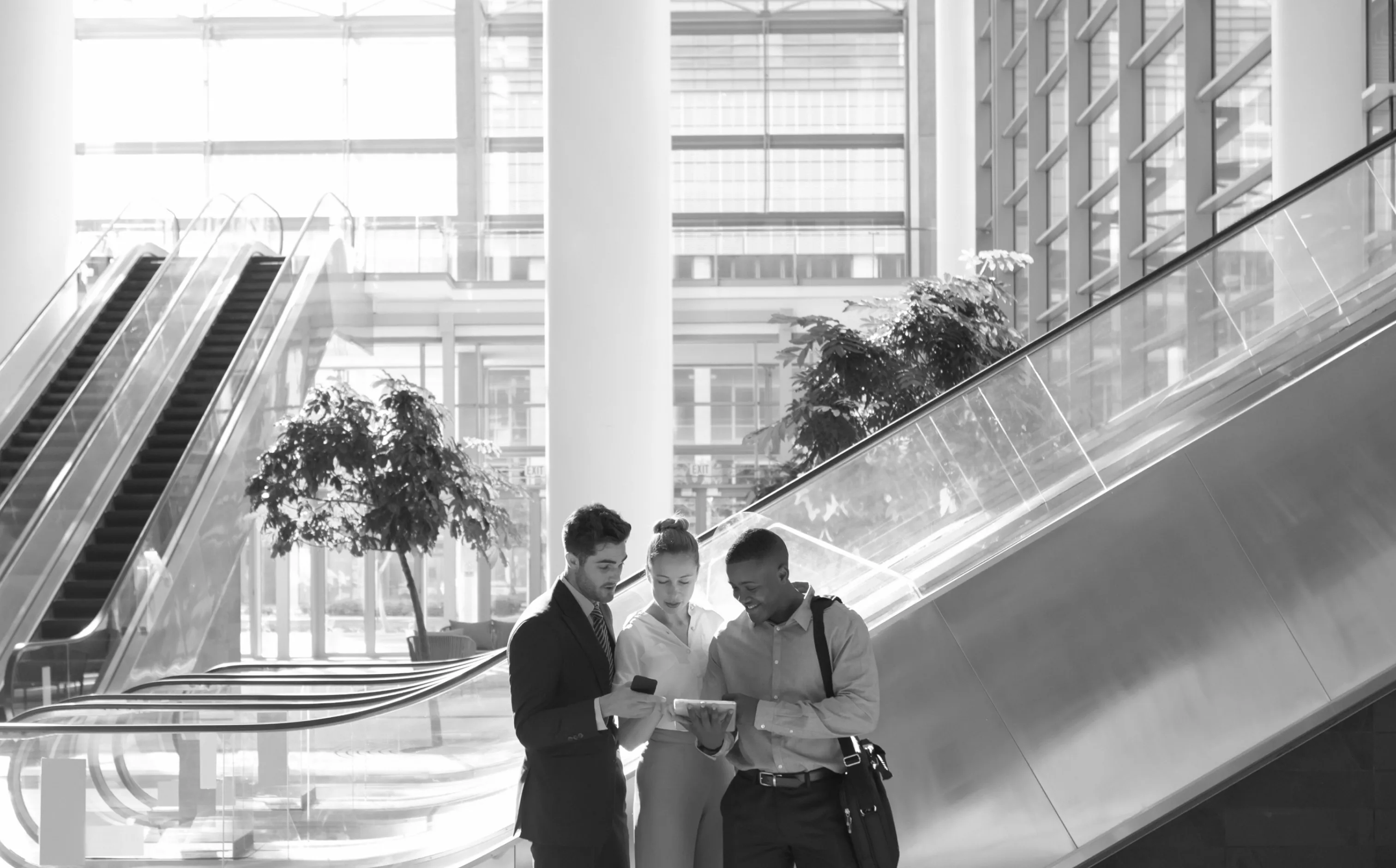 Three business professionals standing together and looking at a tablet in a modern office building with escalators and large windows.