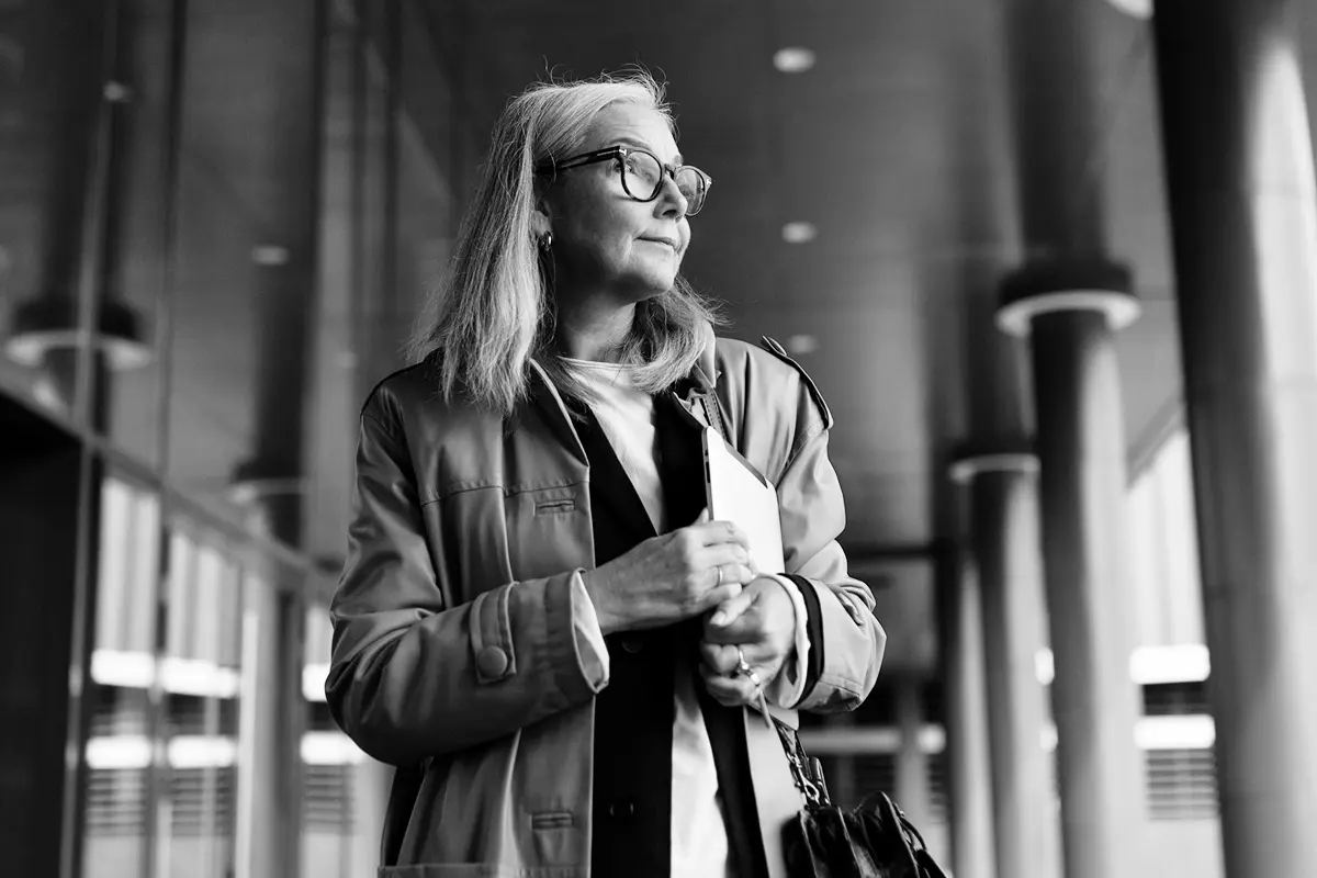Smiling woman with glasses holding a pen and looking at a laptop screen held by a man in a white shirt, seated on a bench indoors.