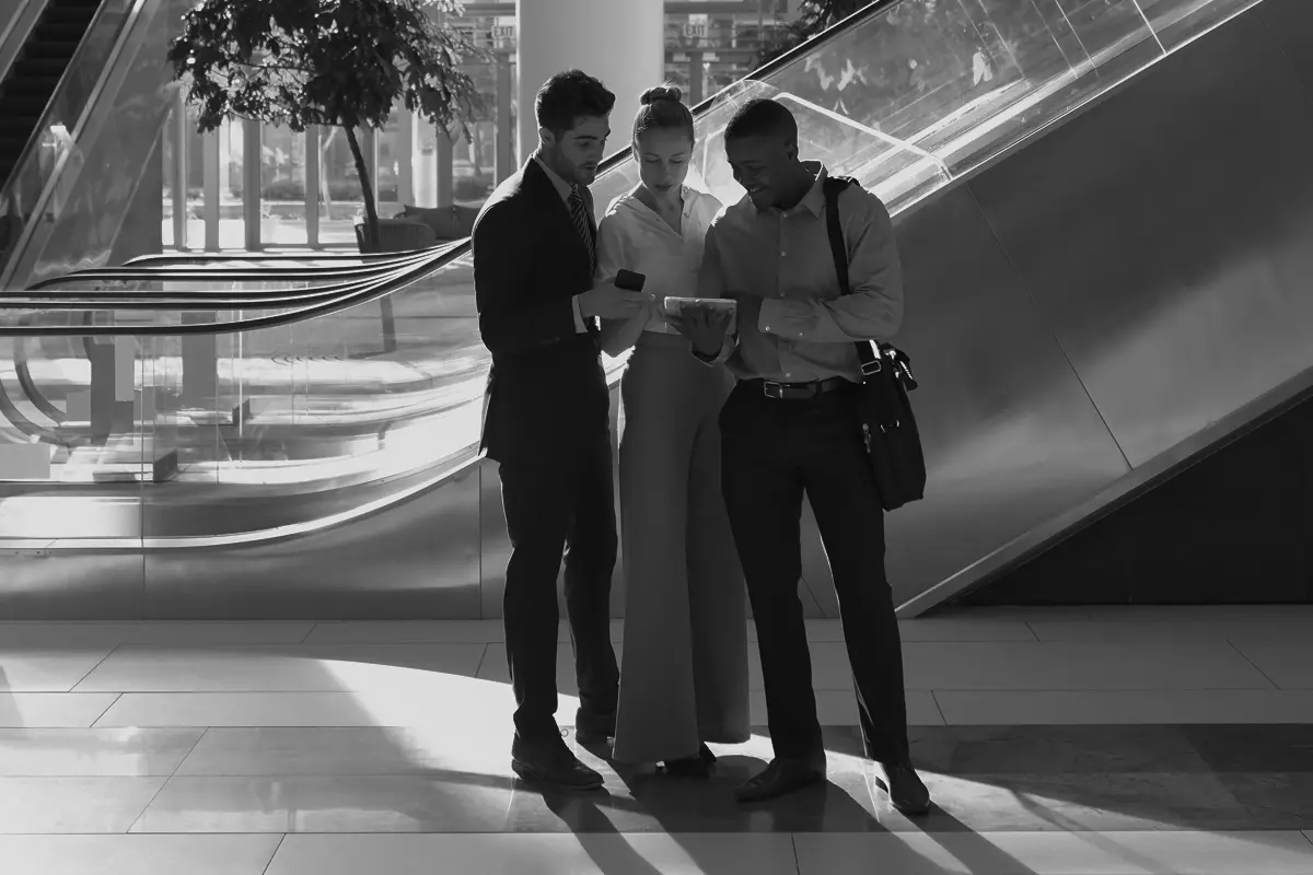Three business professionals standing in a modern building lobby, looking at a tablet together near escalators.
