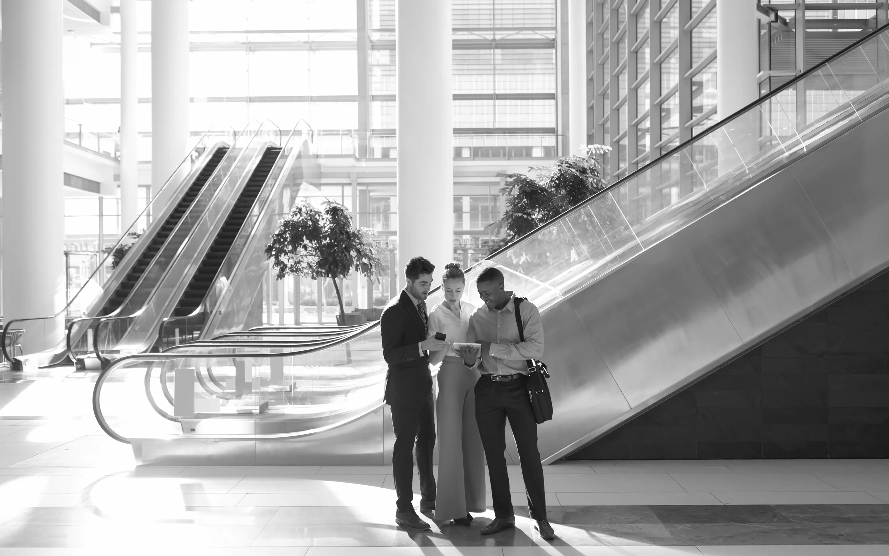 Three business professionals standing together in a modern building lobby, looking at a tablet device.