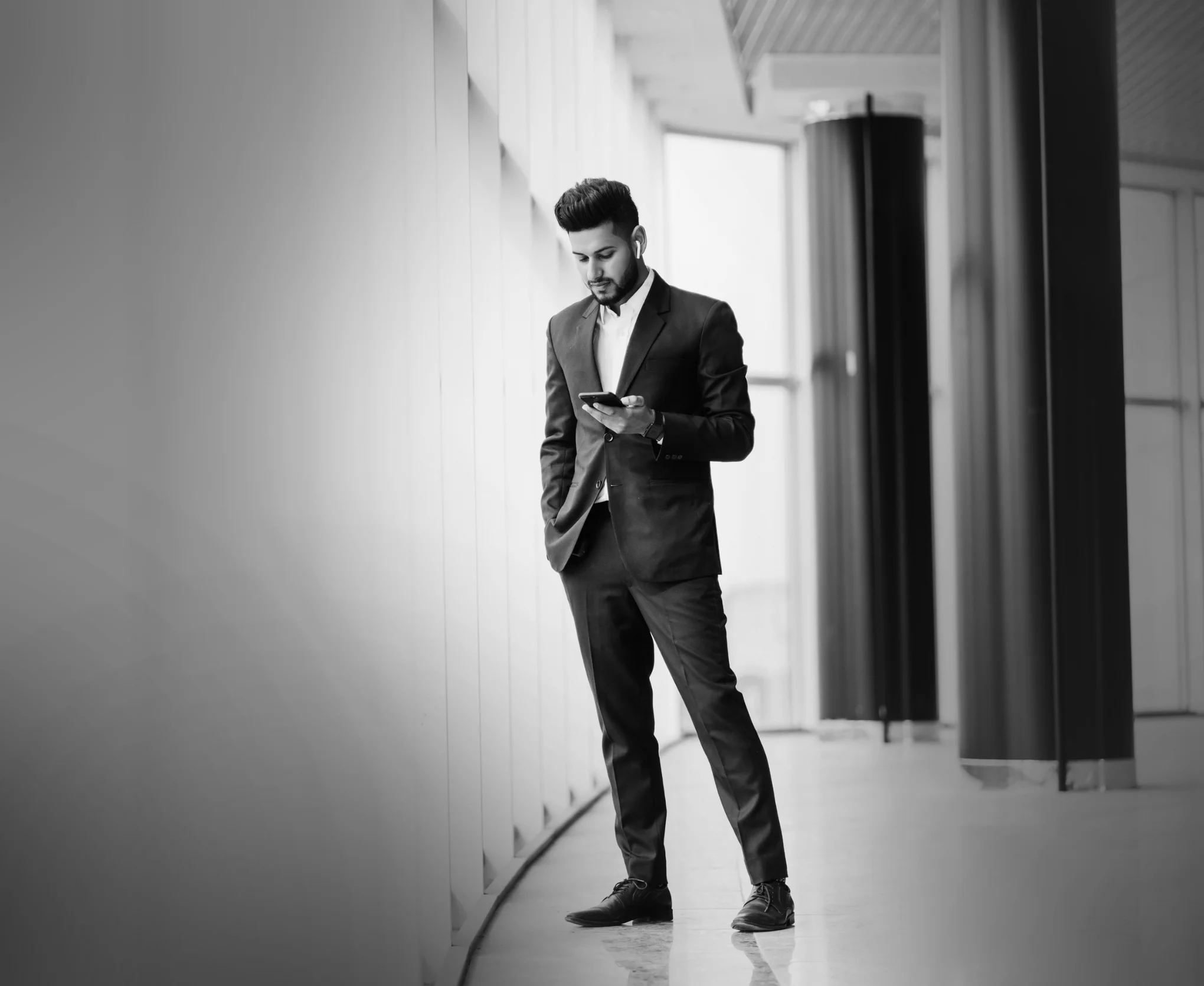 Man in a suit standing by large windows looking at his phone with one hand in pocket.