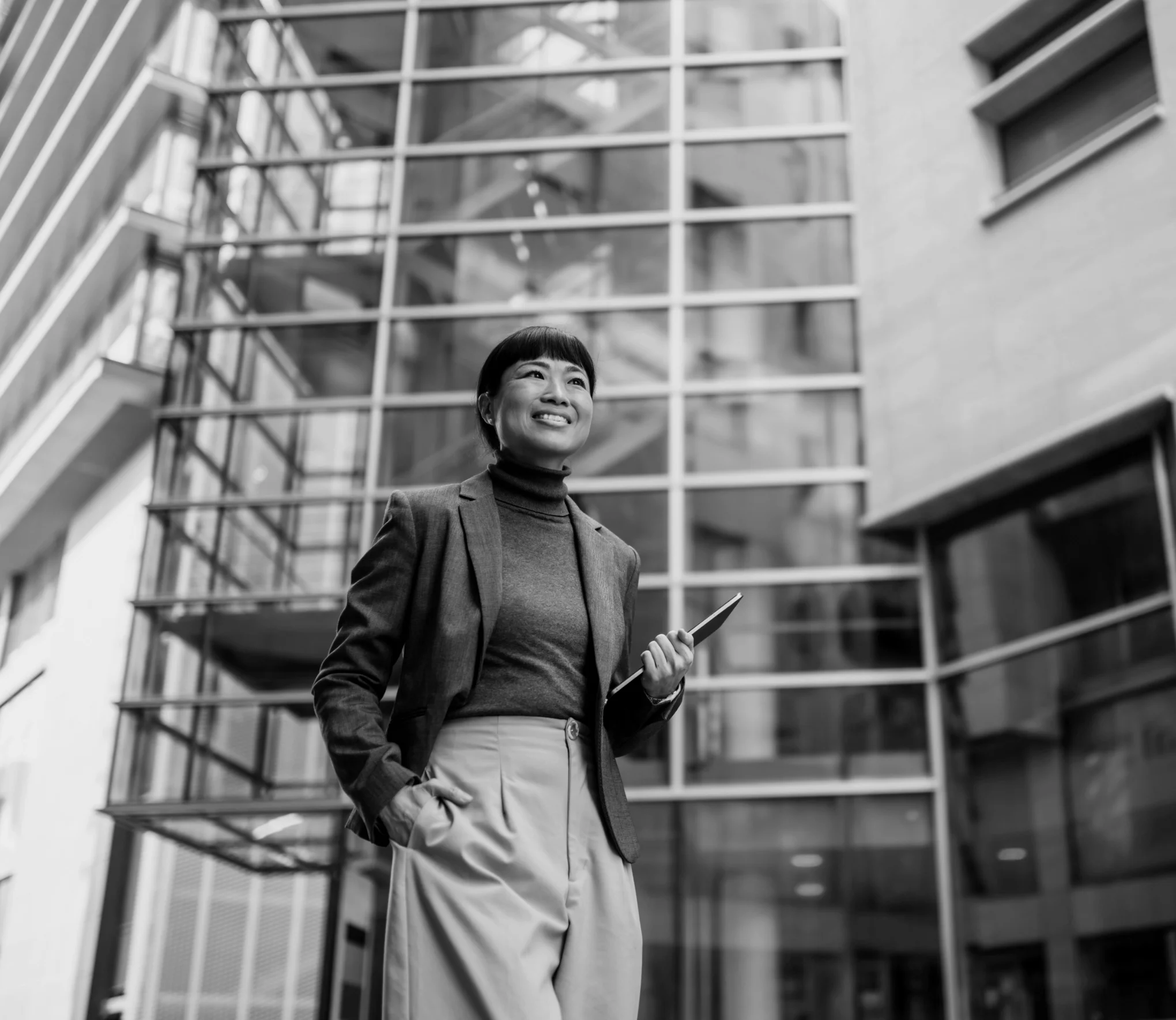 Smiling professional Asian woman holding a tablet, standing in front of a modern glass office building.