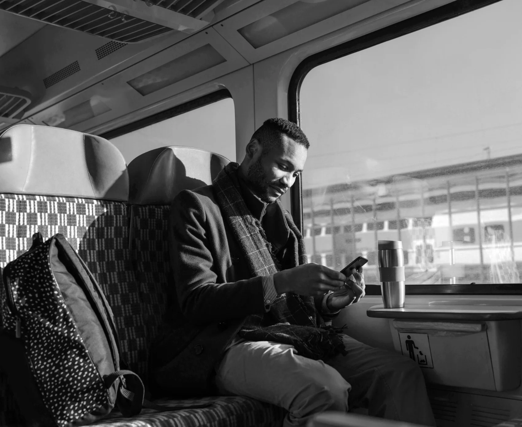 Man sitting on a train seat looking at his smartphone with a travel mug on the table beside him.