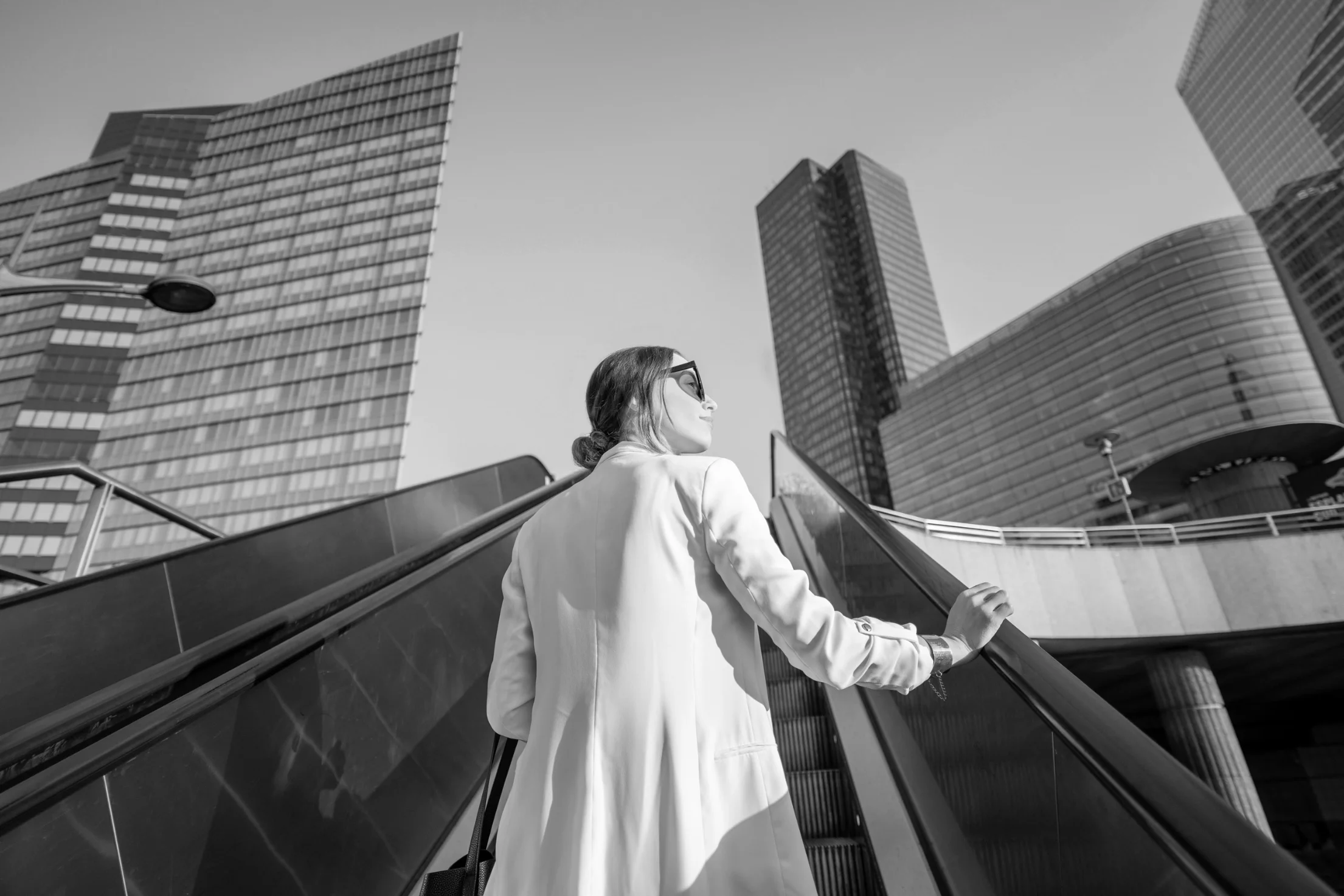 Woman in light coat and sunglasses ascending an outdoor escalator surrounded by modern skyscrapers.