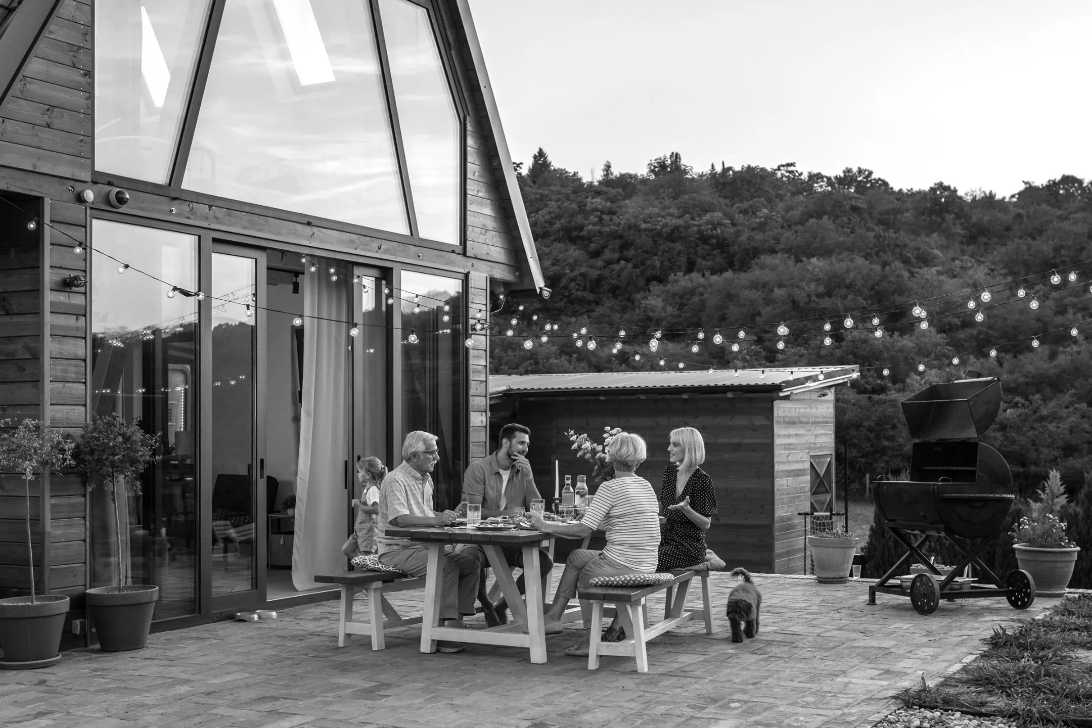 Family of four sitting and chatting at a wooden outdoor table on a patio with string lights overhead and a dog nearby.