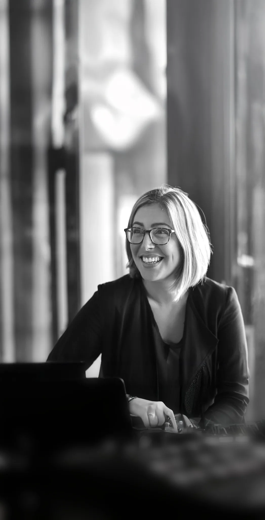 Smiling woman with glasses and shoulder-length hair wearing a dark jacket, looking to the side.