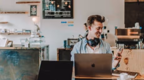 Man smiling at phone while working in a cafe on laptop