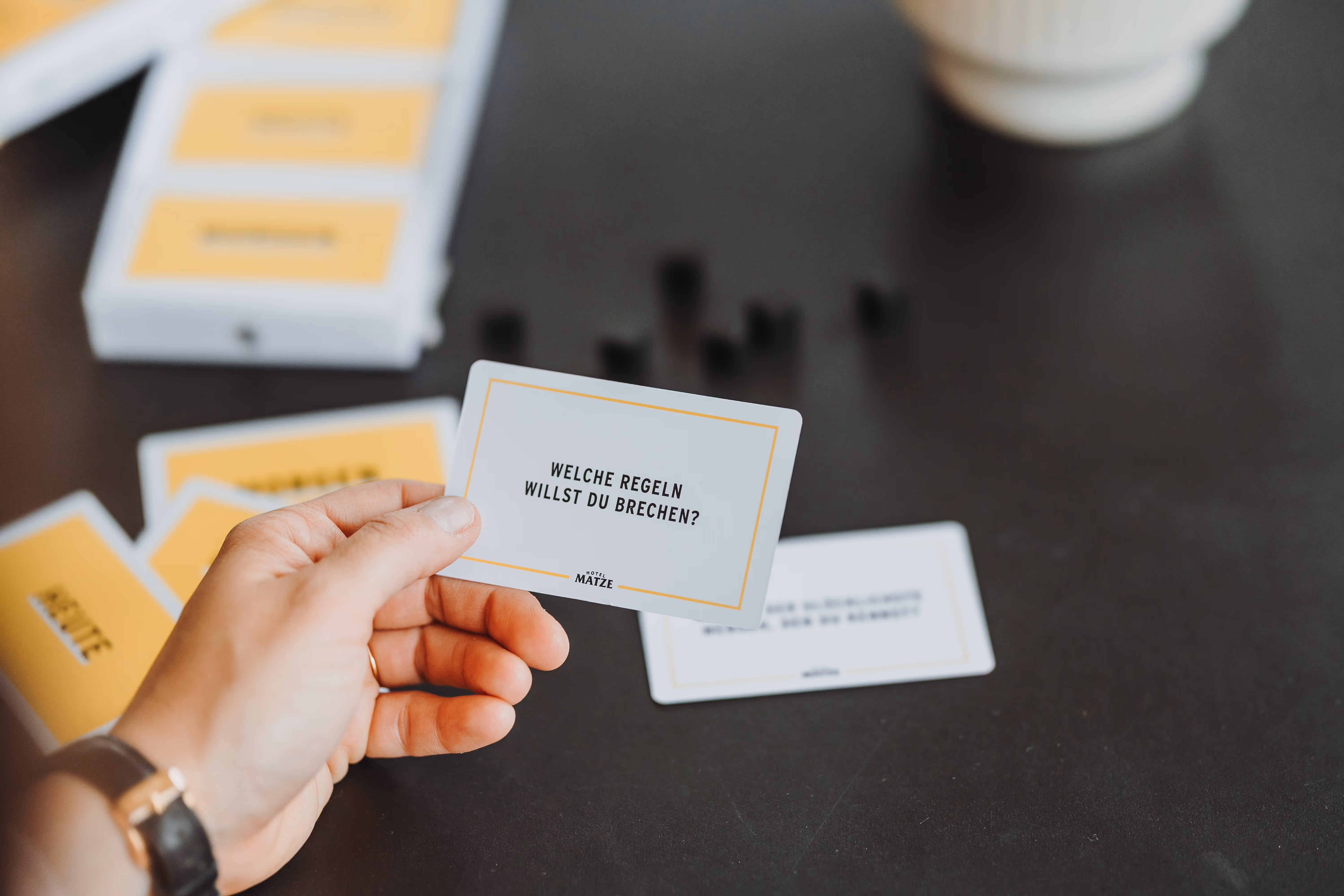 Hand hält Karte mit der Aufschrift „Welche Regeln willst du brechen?“ auf dunklem Tisch mit weiteren Karten und einem Becher im Hintergrund.