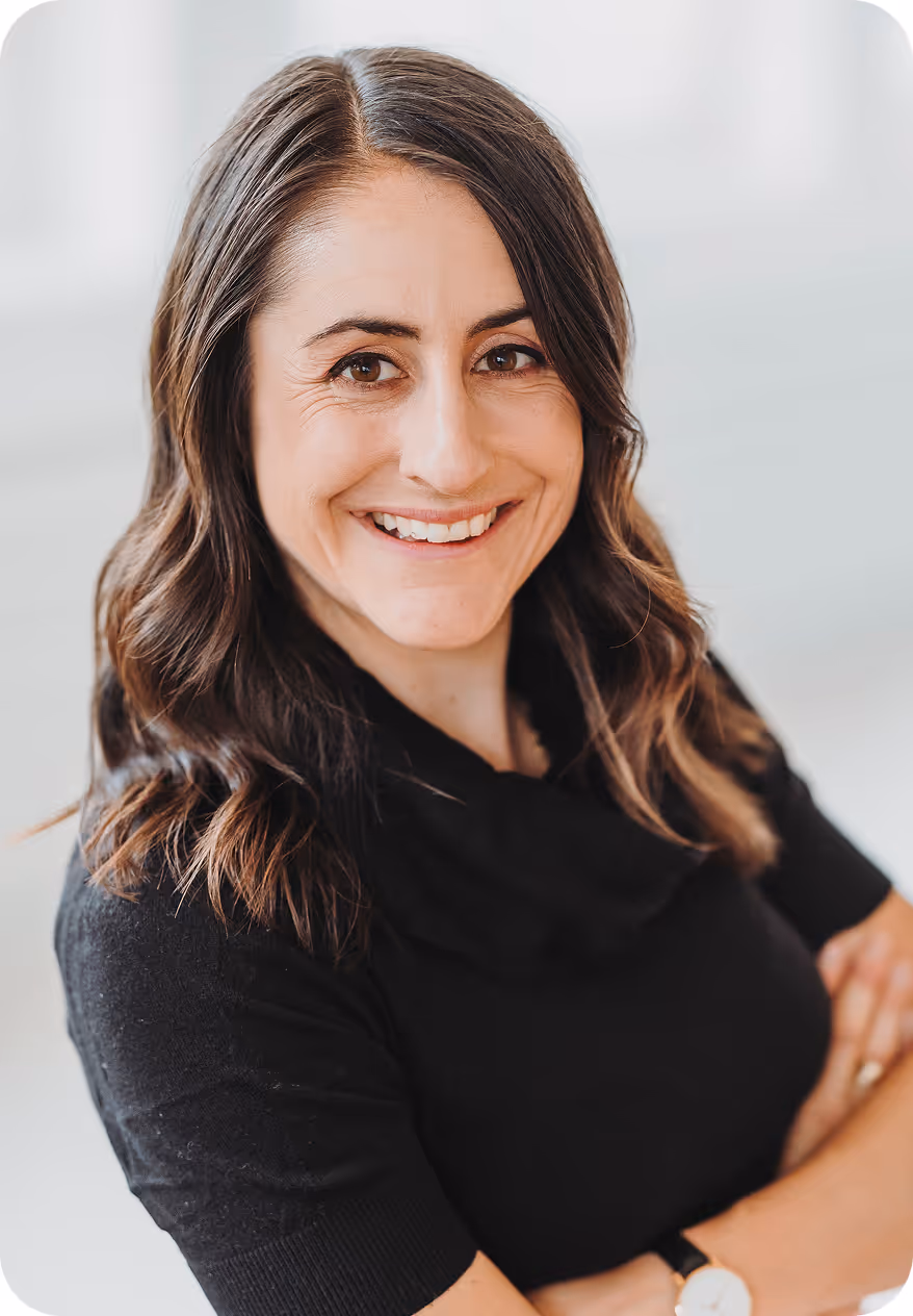 Sandra mit gewellten braunen Haaren in schwarzem Pullover lächelt freundlich mit verschränkten Armen.