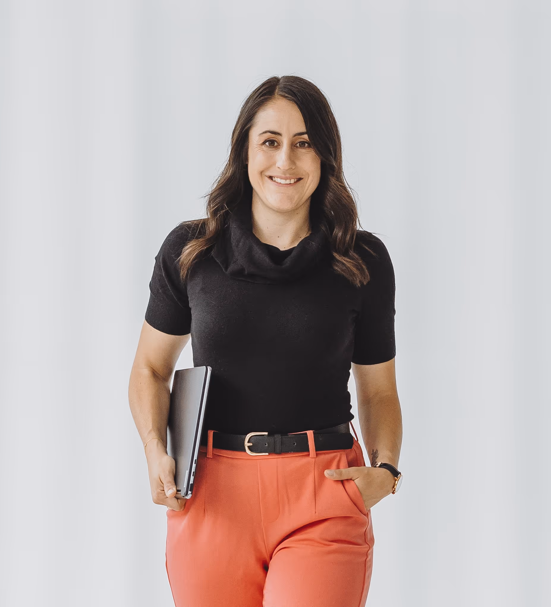 Sandra mit schwarzen Haaren, schwarzem Oberteil und roten Hosen hält einen Laptop, vor hellem Hintergrund.