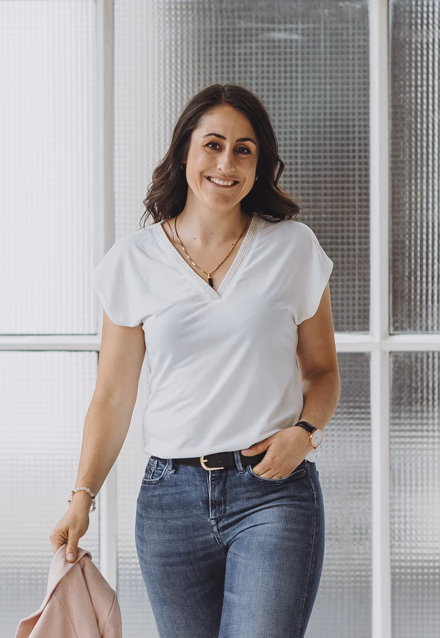 Sandra lächelnd mit lockigem braunem Haar trägt weisses T-Shirt und blaue Jeans, hält rosafarbenes Kleidungsstück in der Hand.