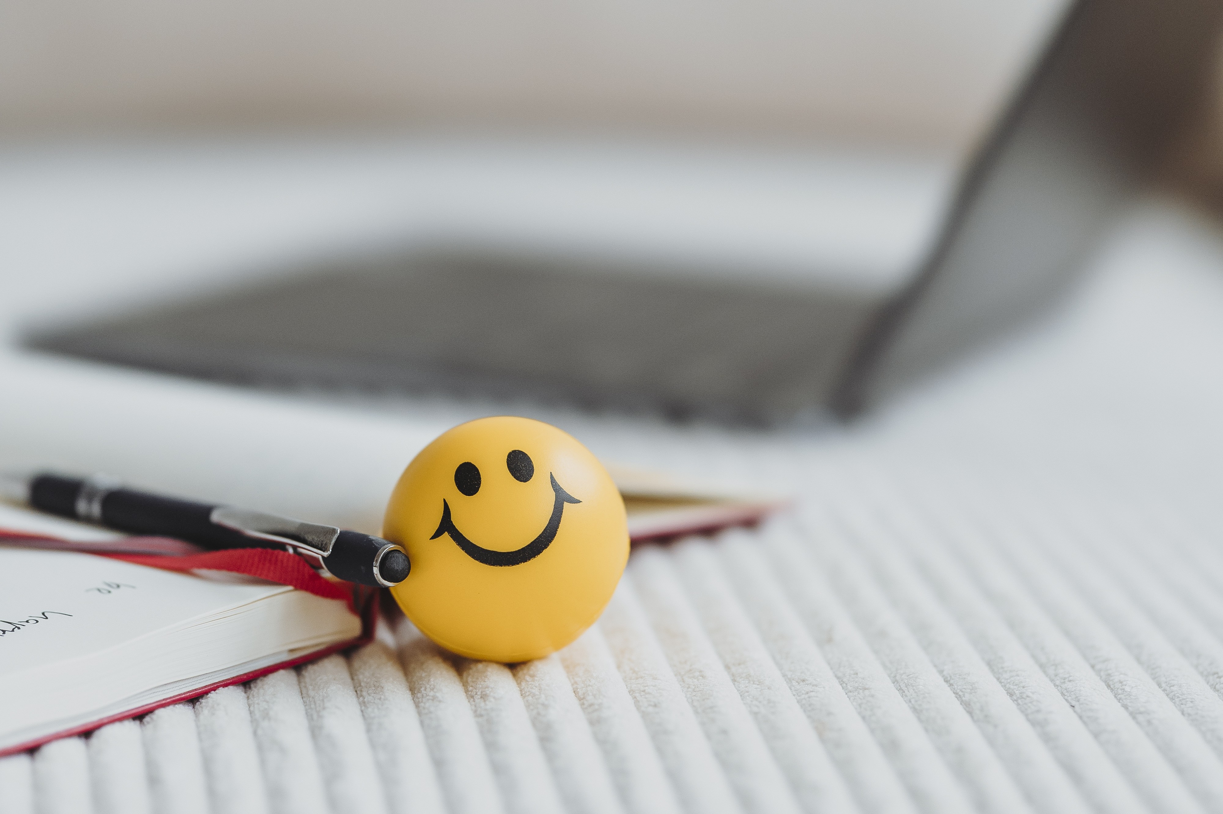 Gelber Stressball mit lächelndem Gesicht neben einem Stift und einem Notizbuch auf einem weißen, strukturierten Stoff.