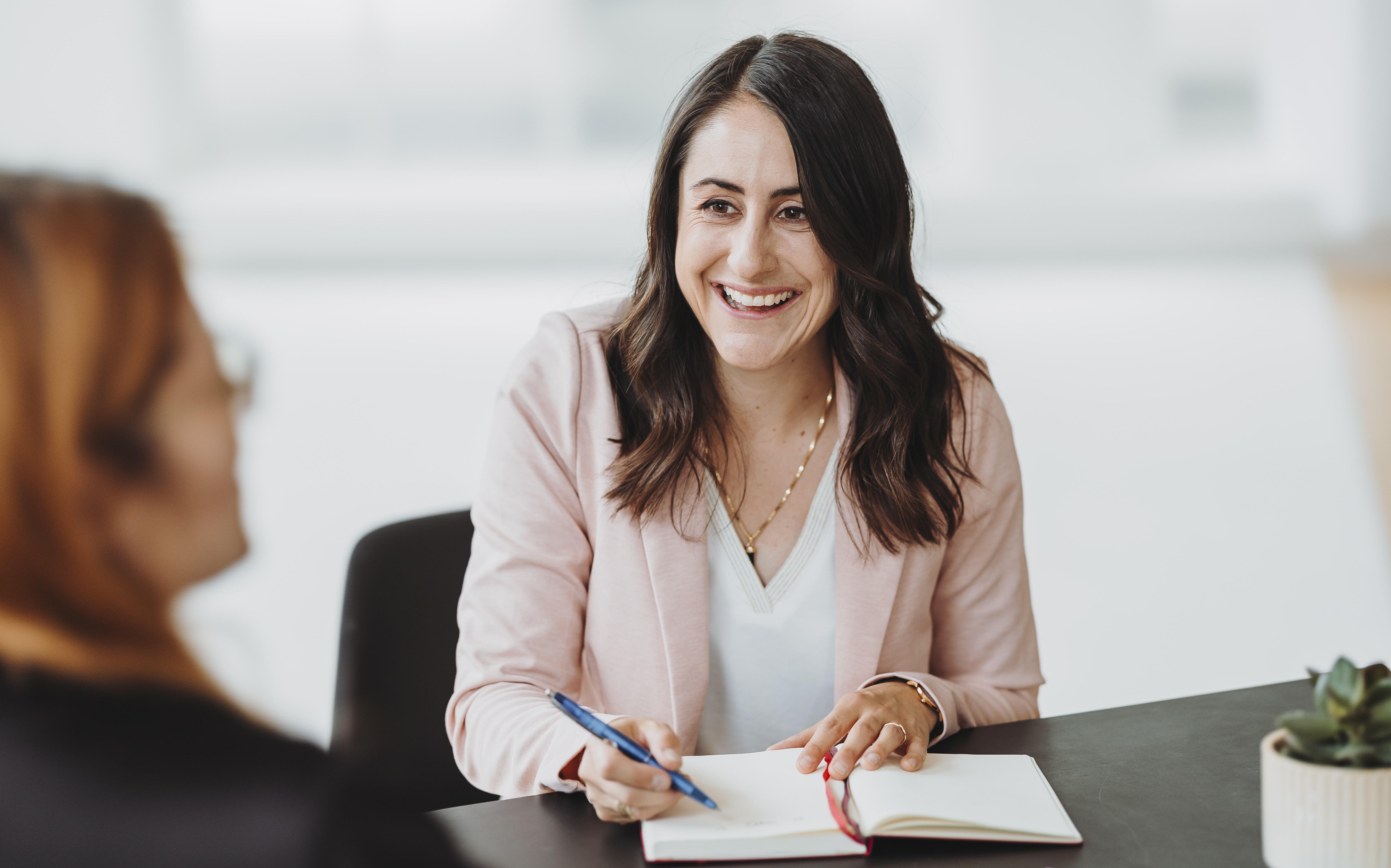 KMU Interims Coach Sandra in rosa Blazer schreibt in einem Notizbuch und spricht mit einer anderen Person.