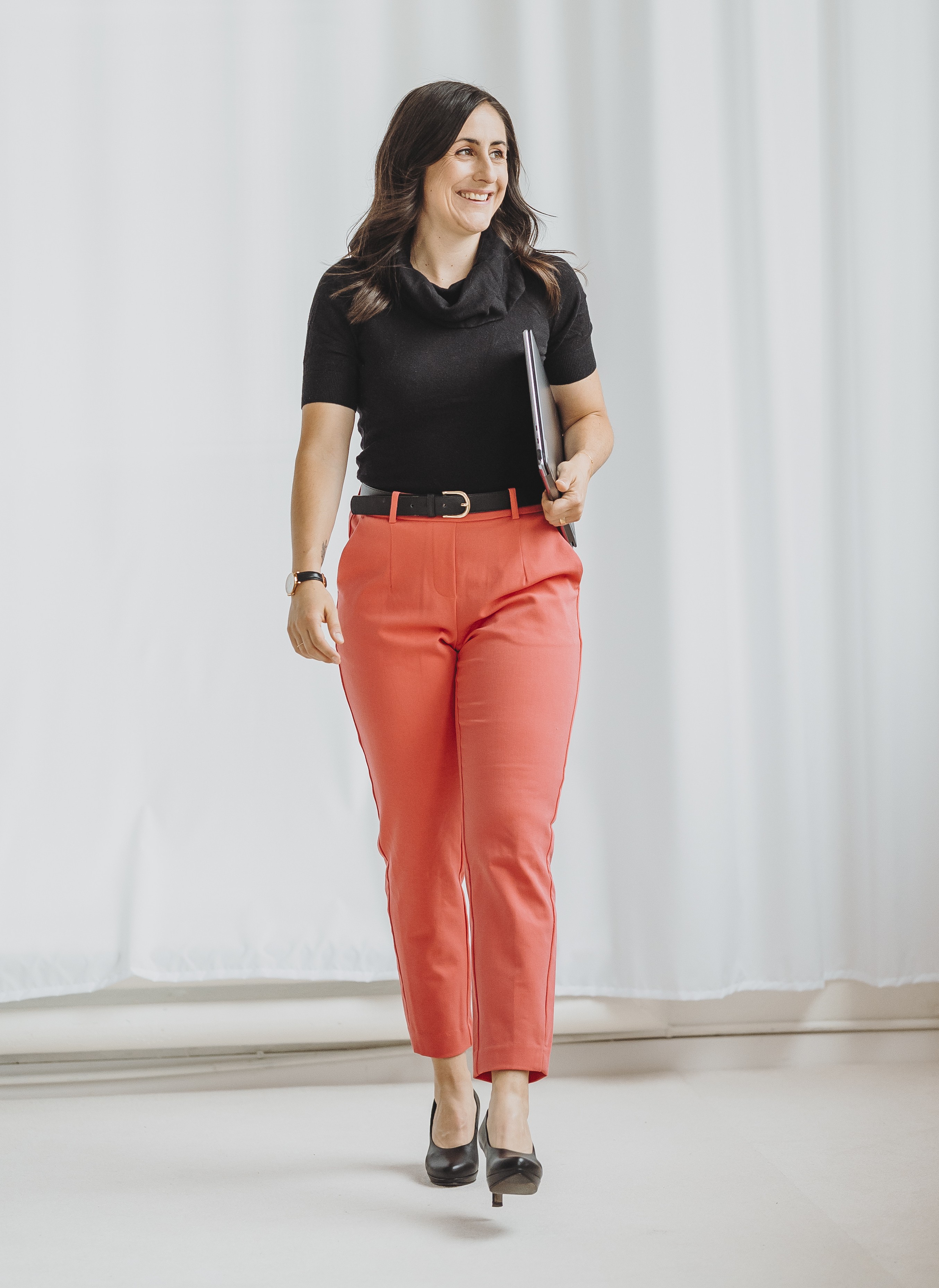 Sandra Wettstein mit schwarzen Oberteil und roten Hosen, die lächelnd einen Laptop hält und in hellem Raum geht.