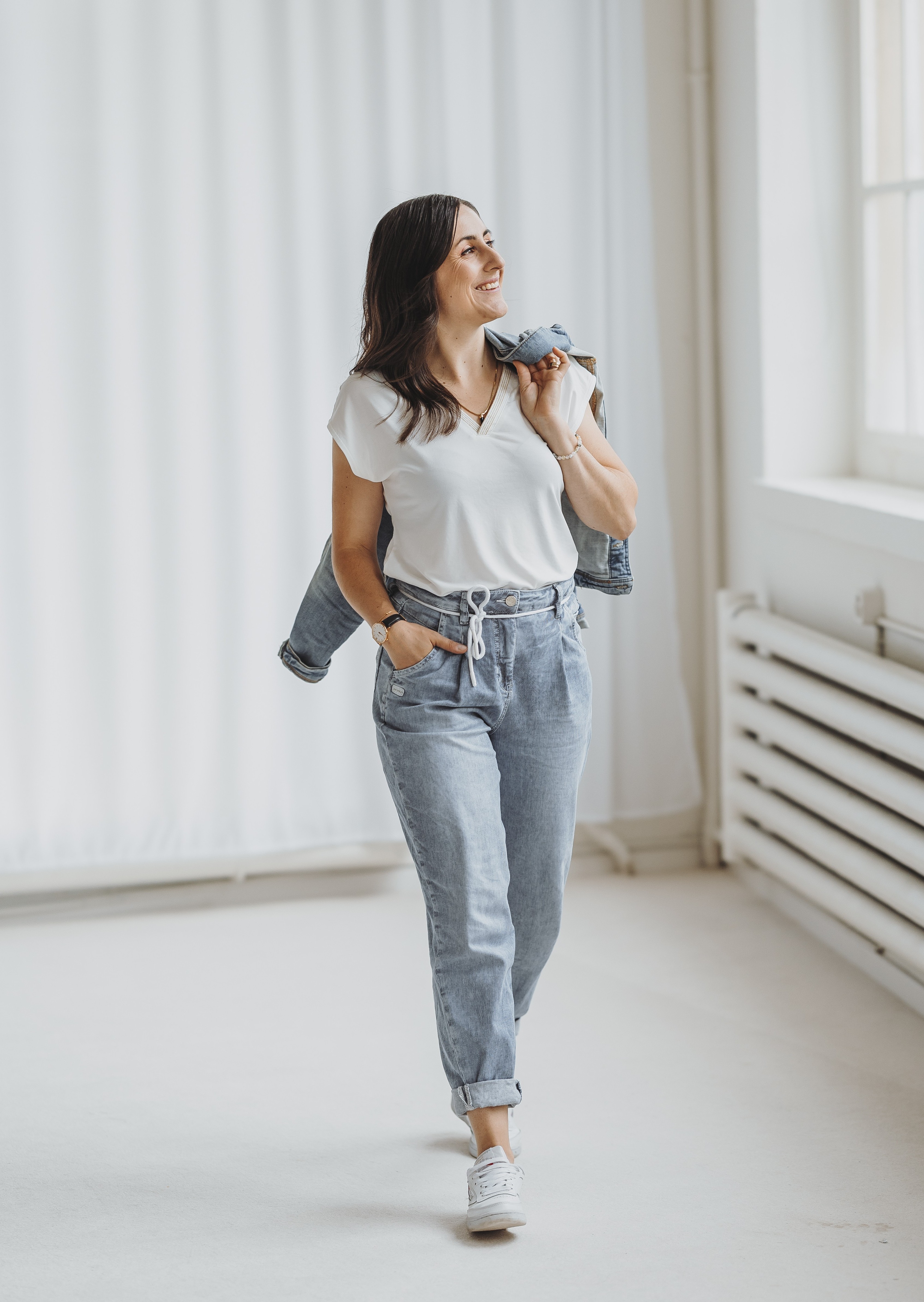 Sandra mit weissem T-Shirt und hellblauen Jeansjacke locker über die Schulter gelegt, die in einem hellen Raum lächelnd geht.
