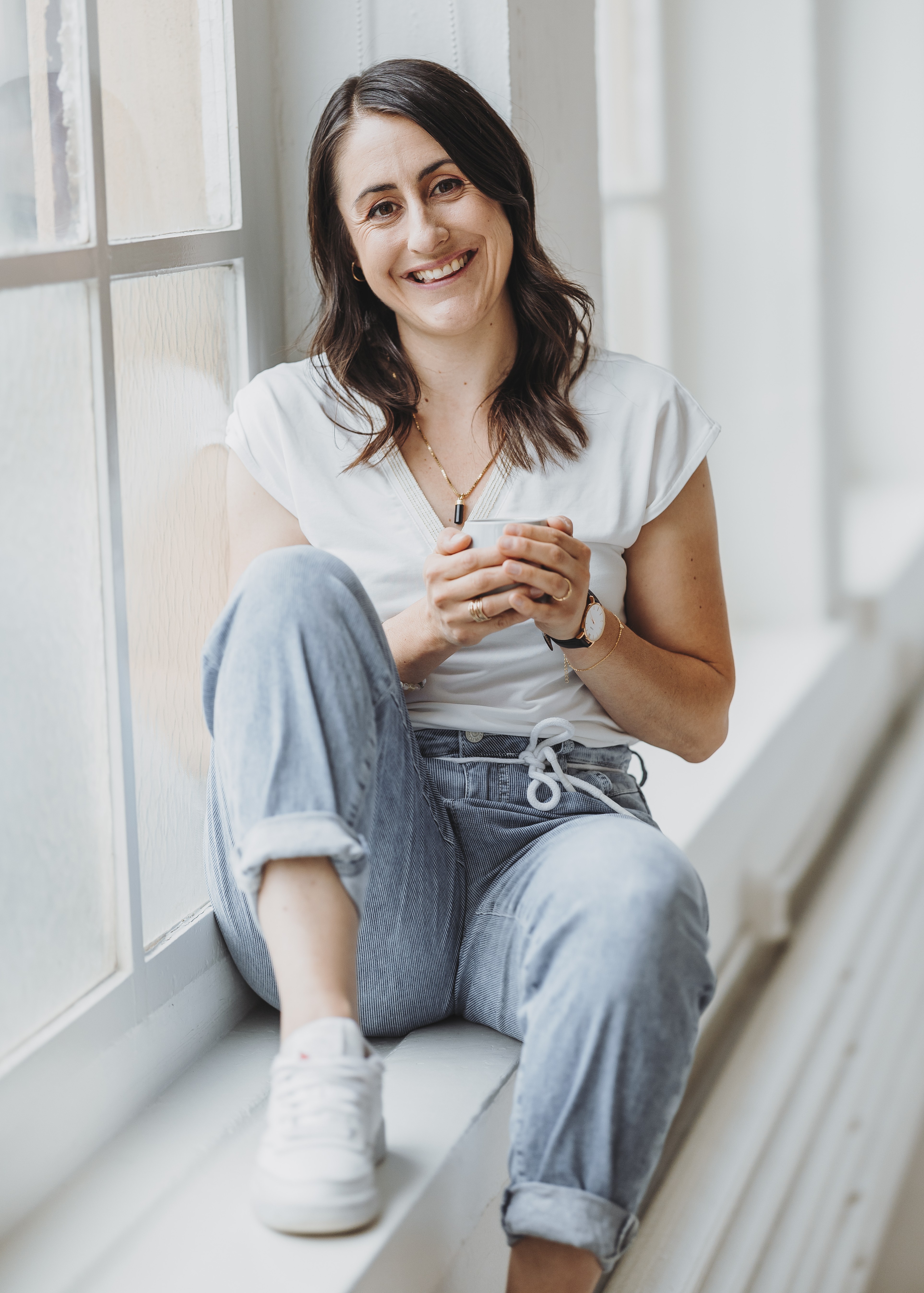 Sandra Wettstein sitzt entspannt auf einem Fenstersims, trägt weiße Turnschuhe, blaue Jeans und ein weisses T-Shirt und hält eine Tasse in den Händen.