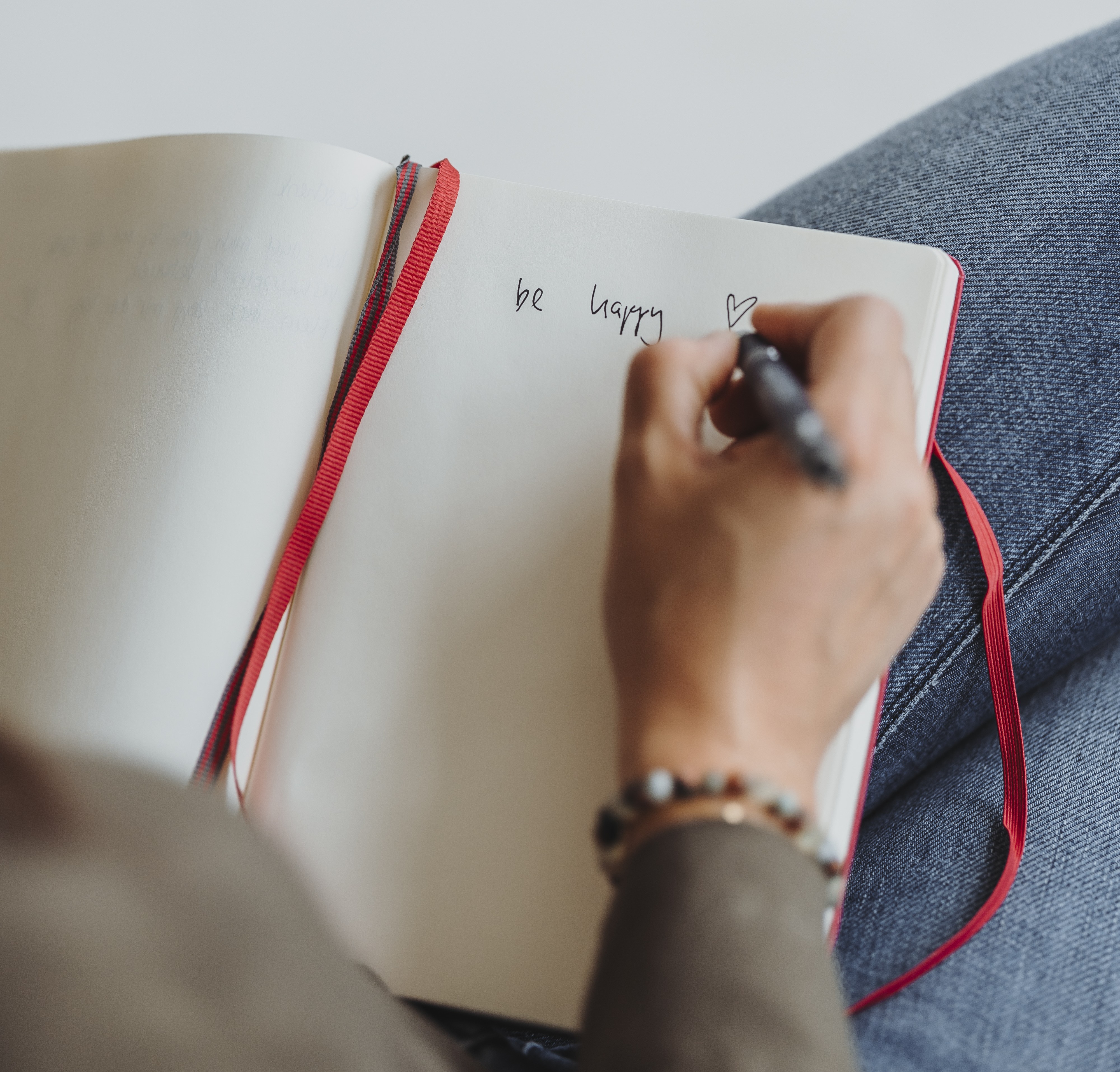 Person schreibt mit einem schwarzen Stift in ein Notizbuch, auf der Seite steht 'be happy' mit einem Herzsymbol.