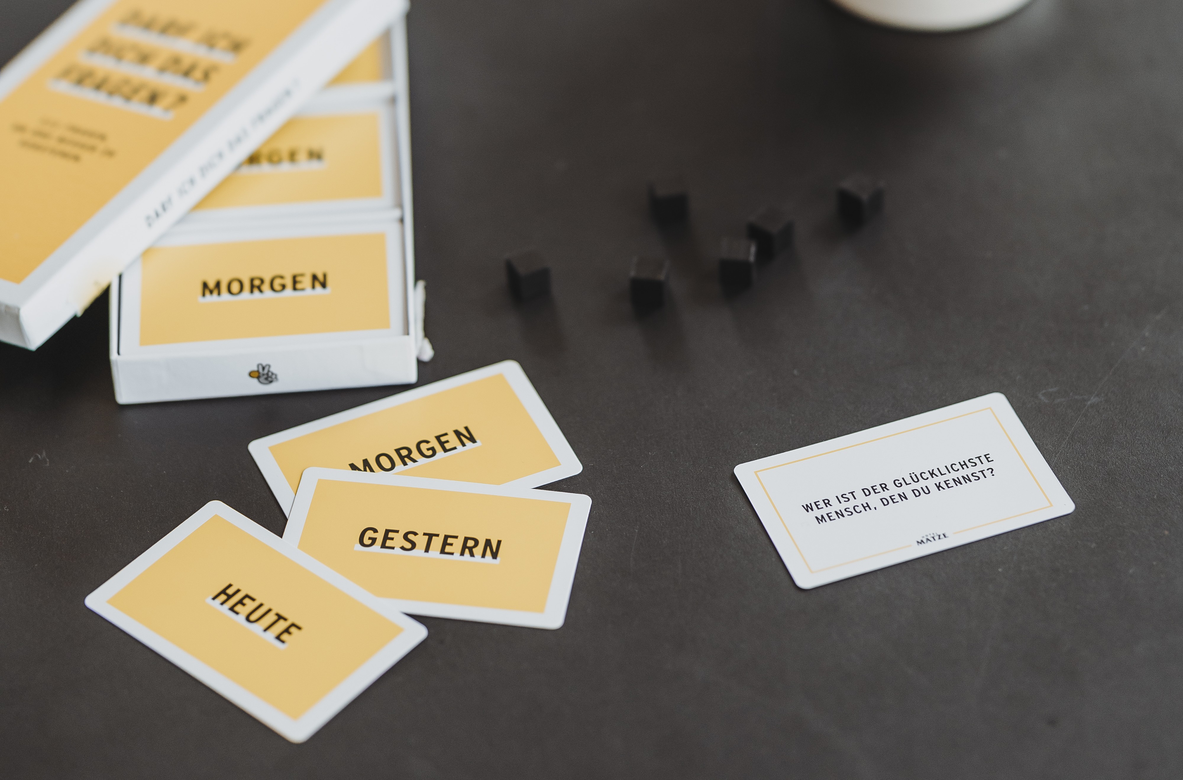 Gelbe Spielkarten mit den Aufschriften Heute, Gestern, Morgen und eine weiße Karte mit der Frage Wer ist der glücklichste Mensch, den du kennst? liegen auf einem dunklen Tisch neben schwarzen Würfeln.