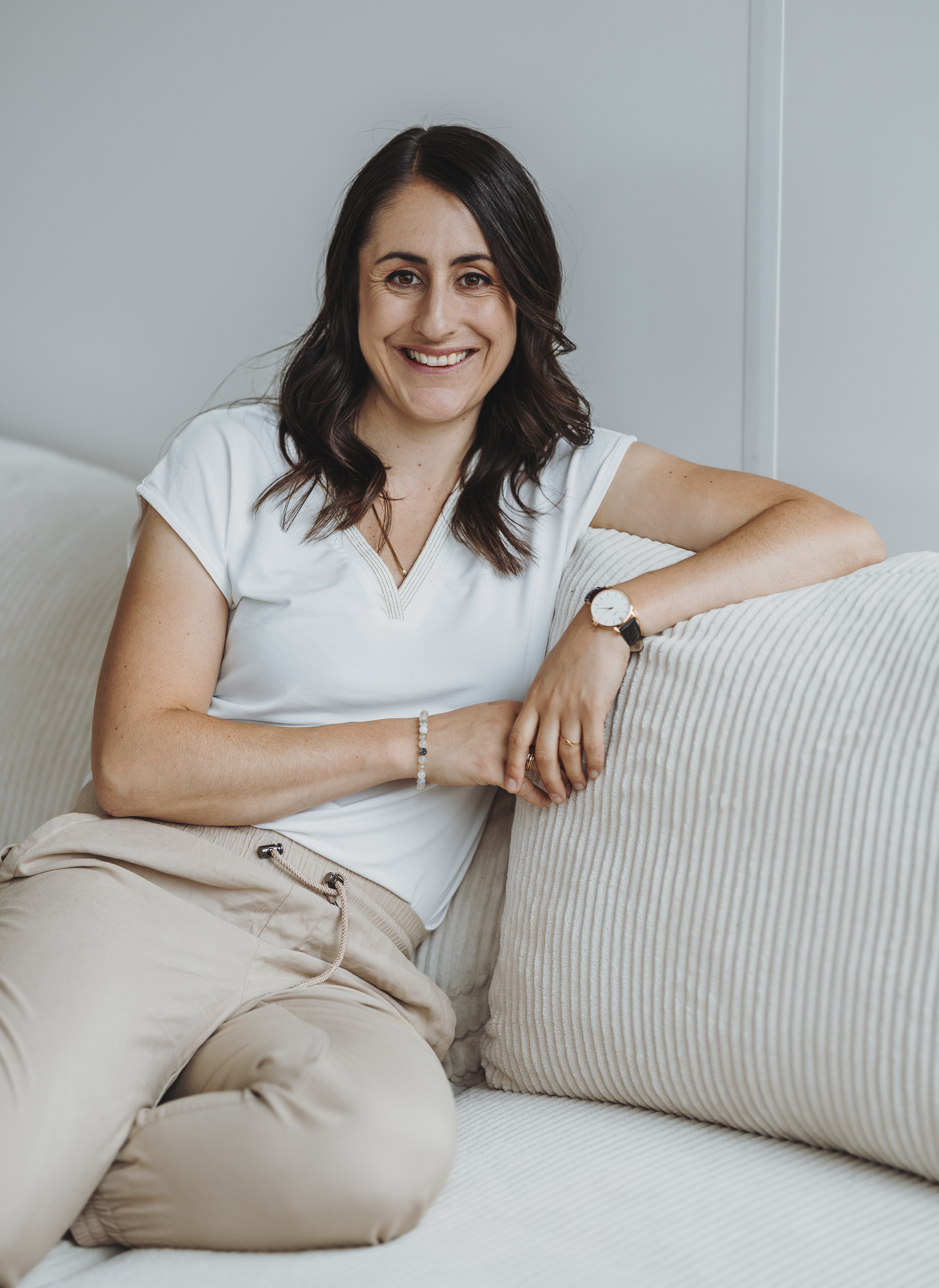 Frau mit dunkelbraunen, schulterlangen Haaren und weißem T-Shirt sitzt entspannt auf einem hellen Sofa und lächelt.