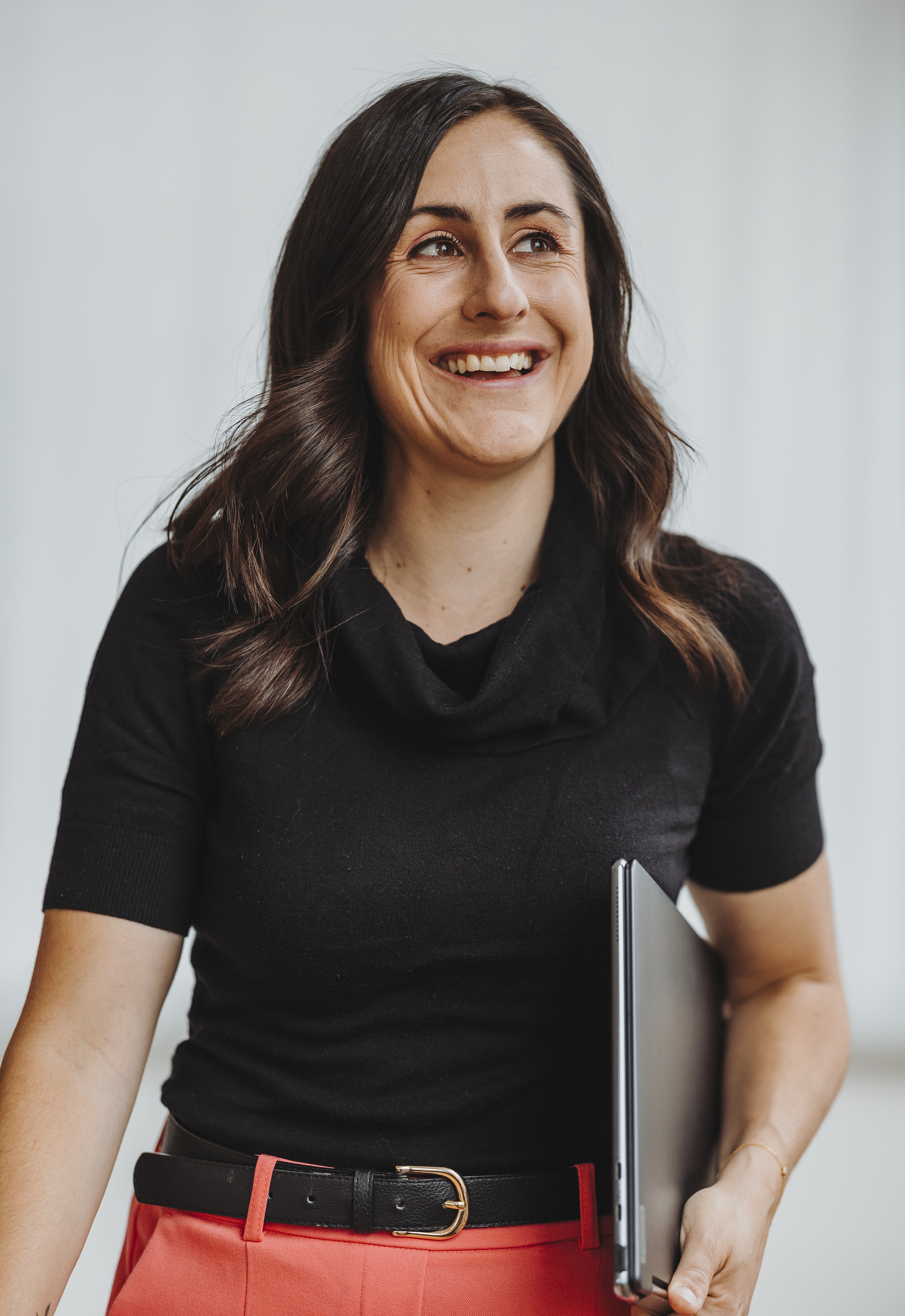 Sandra Wettstein mit schwarzen Haaren, schwarzem Oberteil und roter Hose, die lächelnd einen Laptop hält.