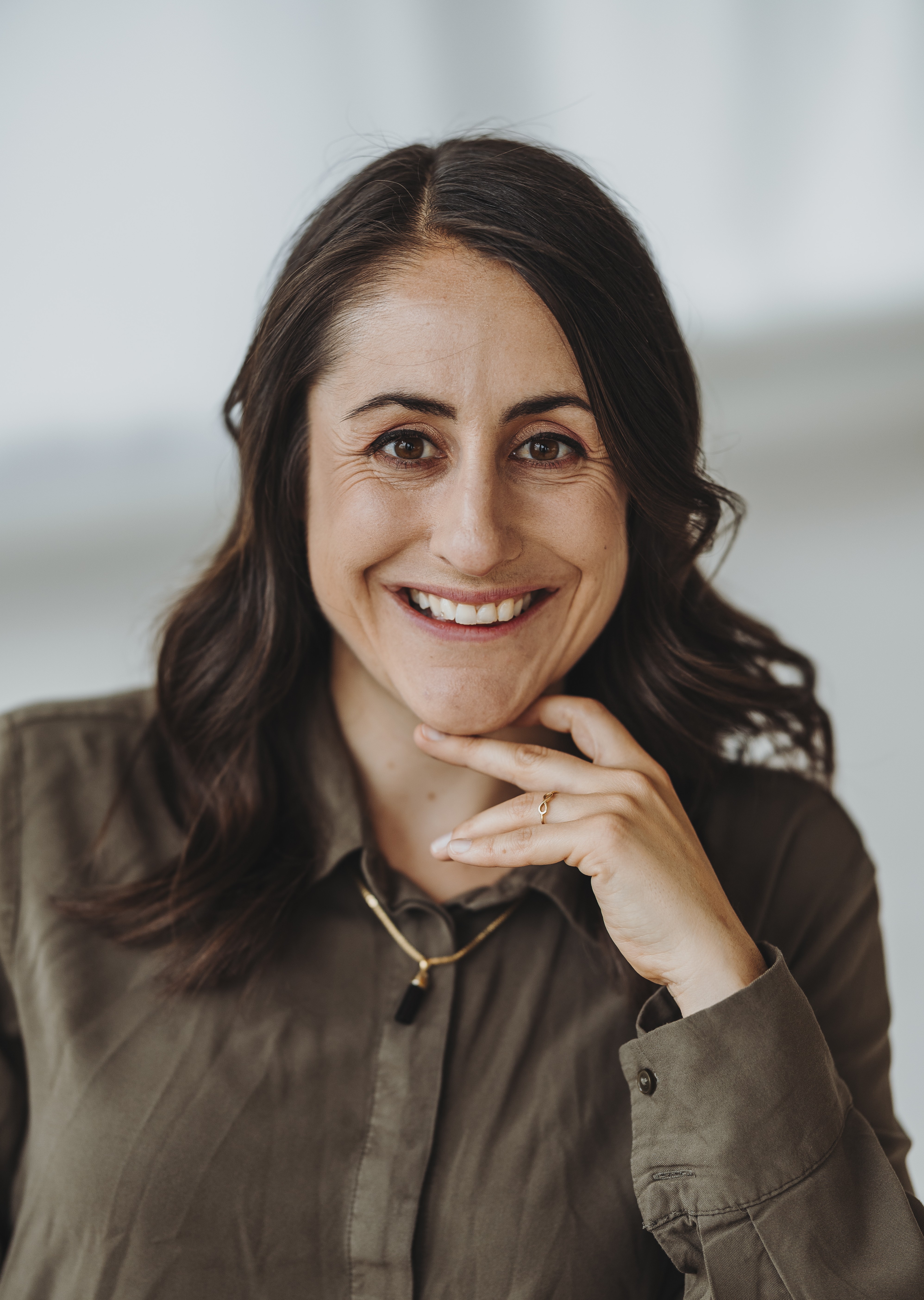 Lächelnde Sandra Wettstein mit braunen Haaren, die ihr Kinn mit der Hand berührt, trägt olivgrünes Hemd und goldene Halskette.