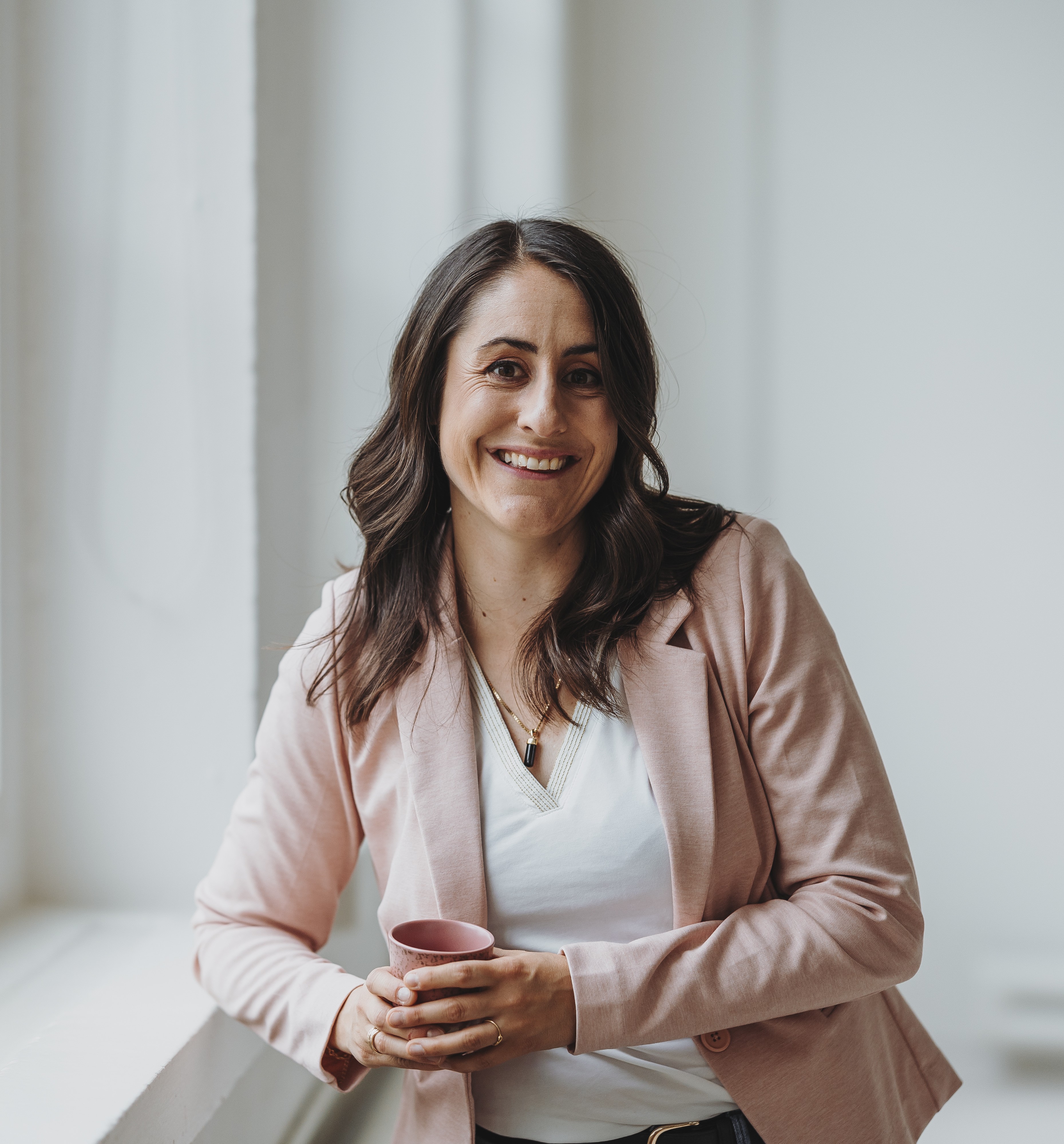 Lächelnde Sandra mit langen braunen Haaren trägt ein rosa Sakko und hält eine rosa Tasse.