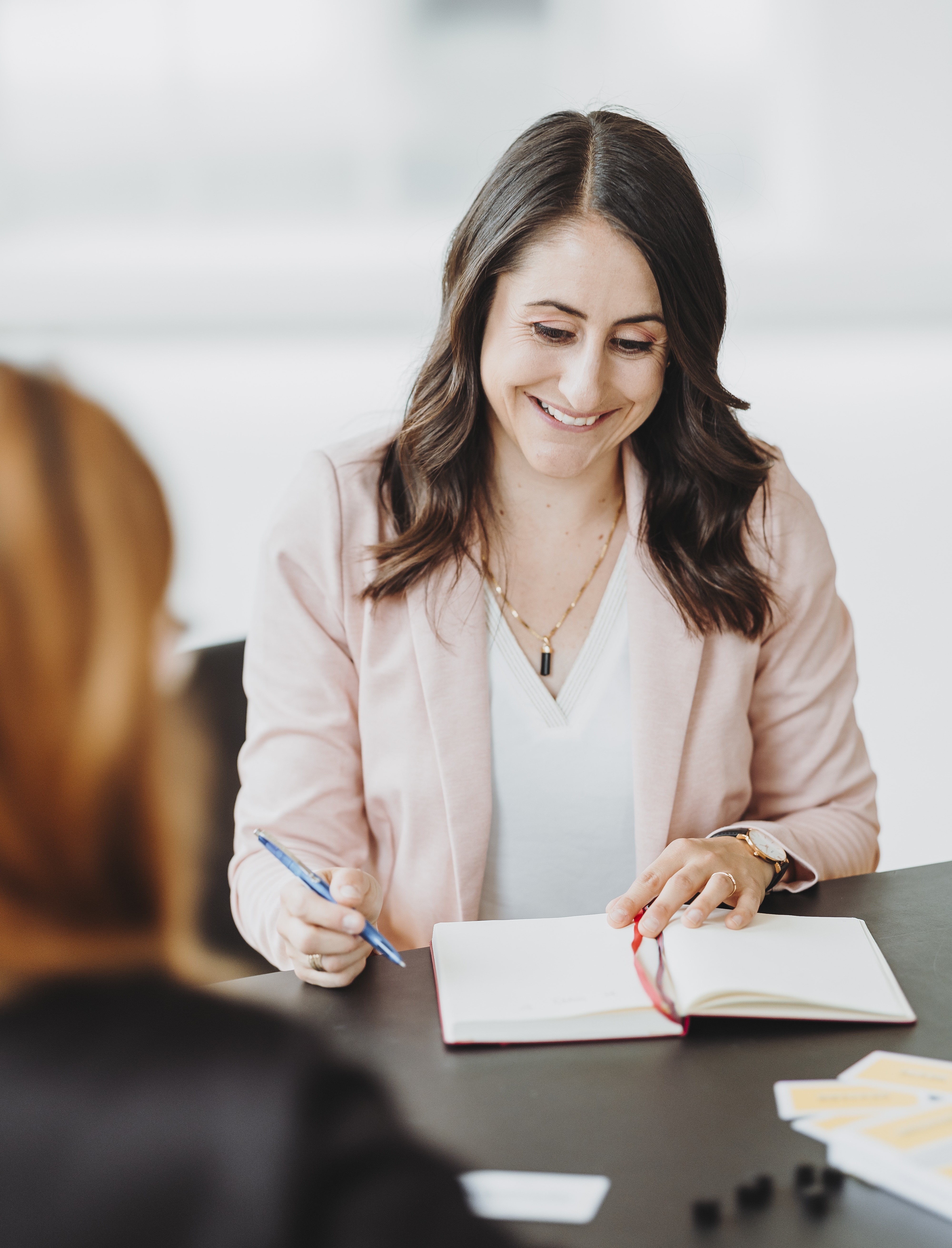 Sandra Wettstein in rosa Blazer, die an einem Tisch sitzt, mit einem offenen Notizbuch und Stift in der Hand.