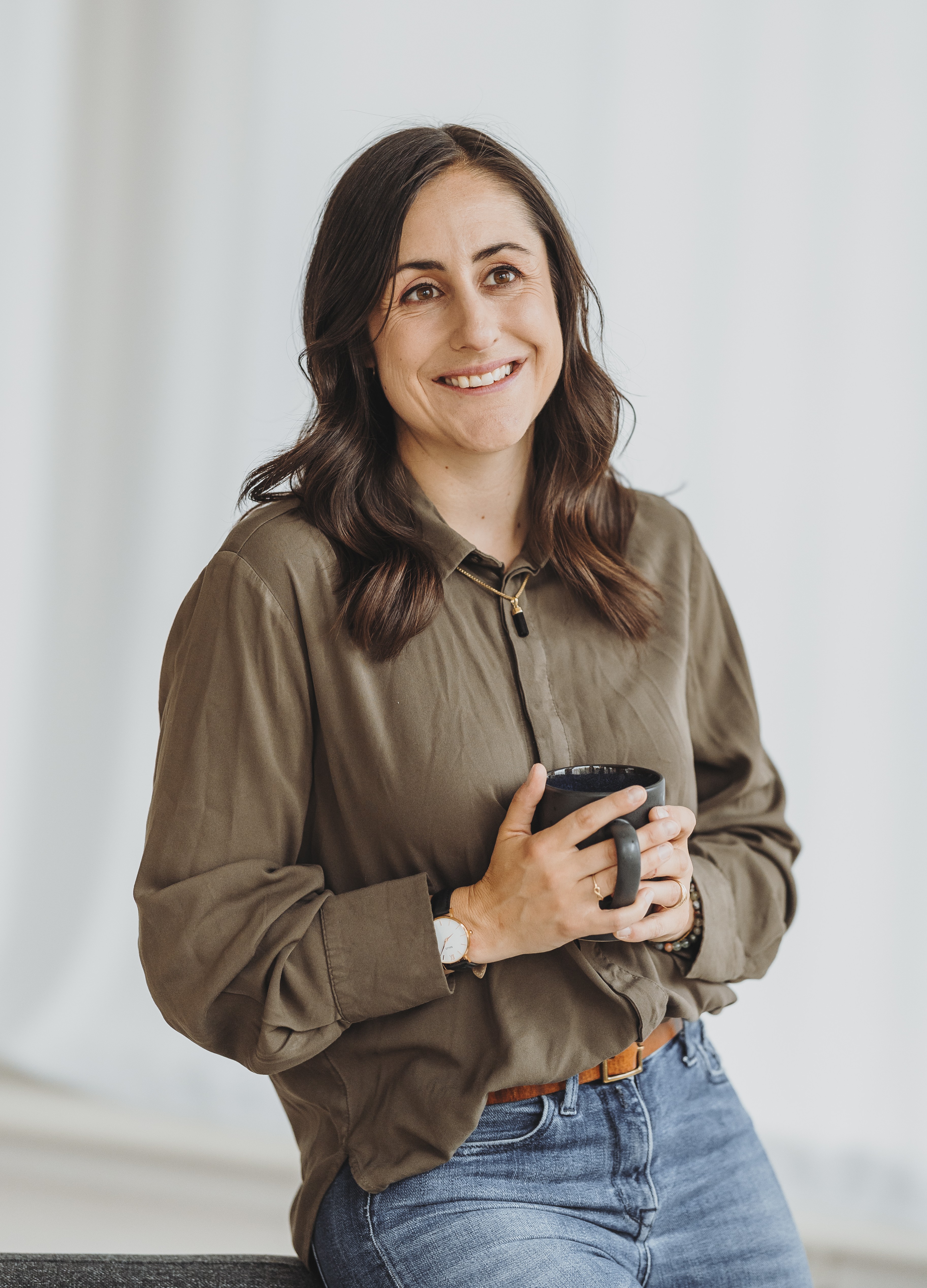 Sandra Wettstein mit langen braunen Haaren lächelt und hält eine schwarze Kaffeetasse, trägt eine olivgrüne Bluse und blaue Jeans.