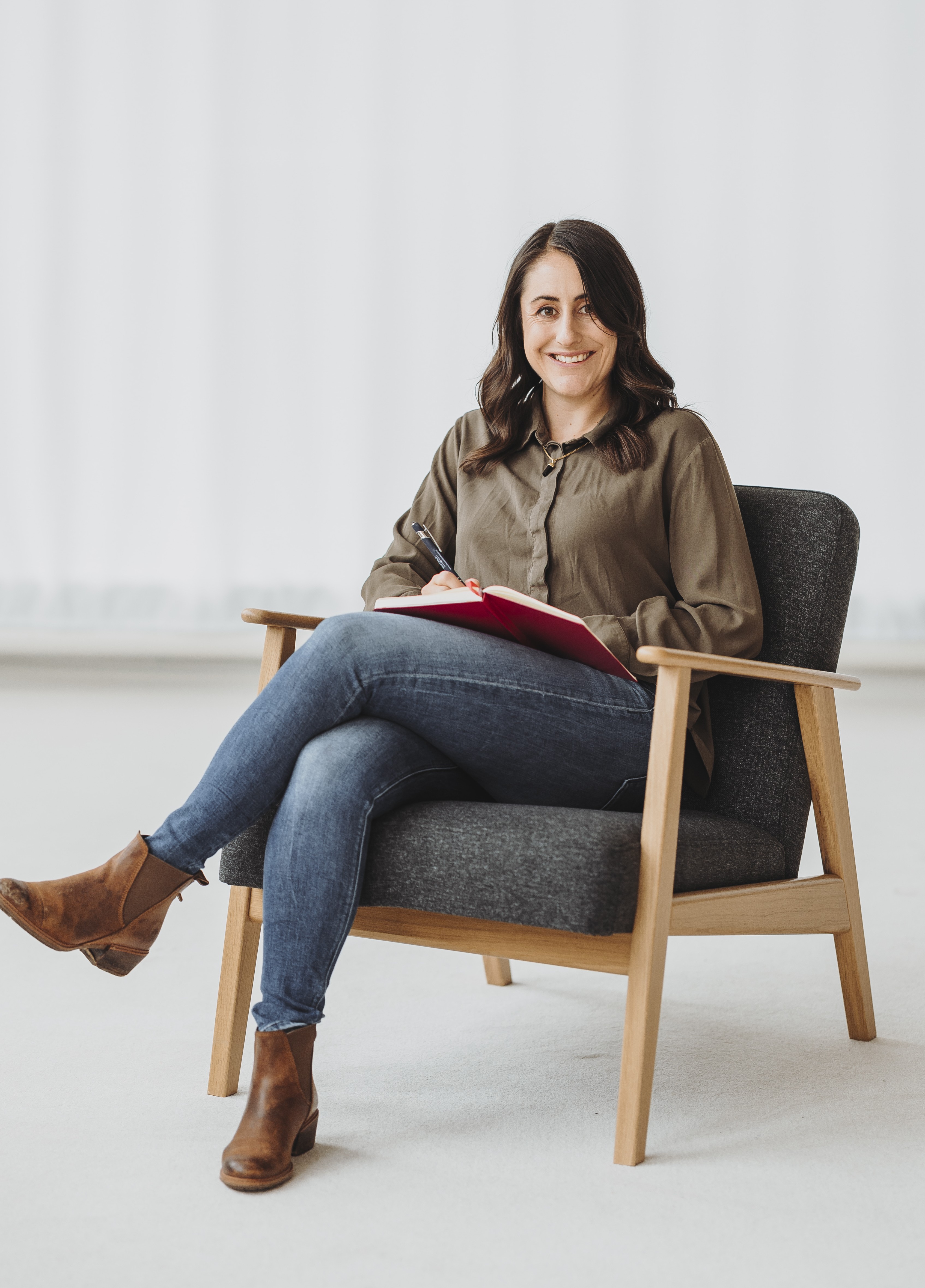 Sandra Wettstein in brauner Bluse und Jeans sitzt auf einem grauen Sessel, schreibt in ein rotes Notizbuch und lächelt.