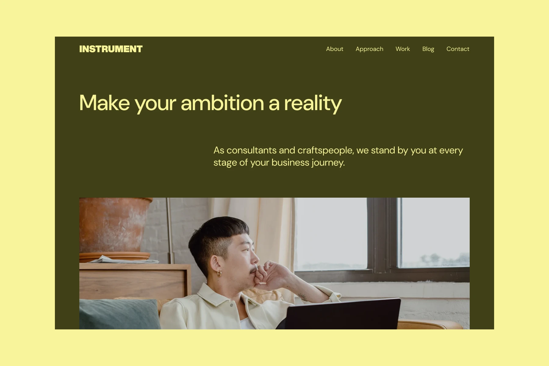 A man sitting on a couch with a laptop, looking thoughtfully out a window in a cozy room.