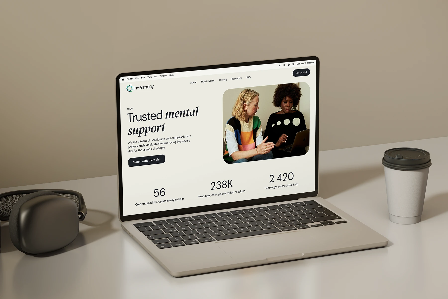 Laptop on a white desk displaying a mental health support website with statistics, a call to match with a therapist, and a photo of two women discussing over a laptop; black headphones and a takeaway coffee cup are beside the laptop.