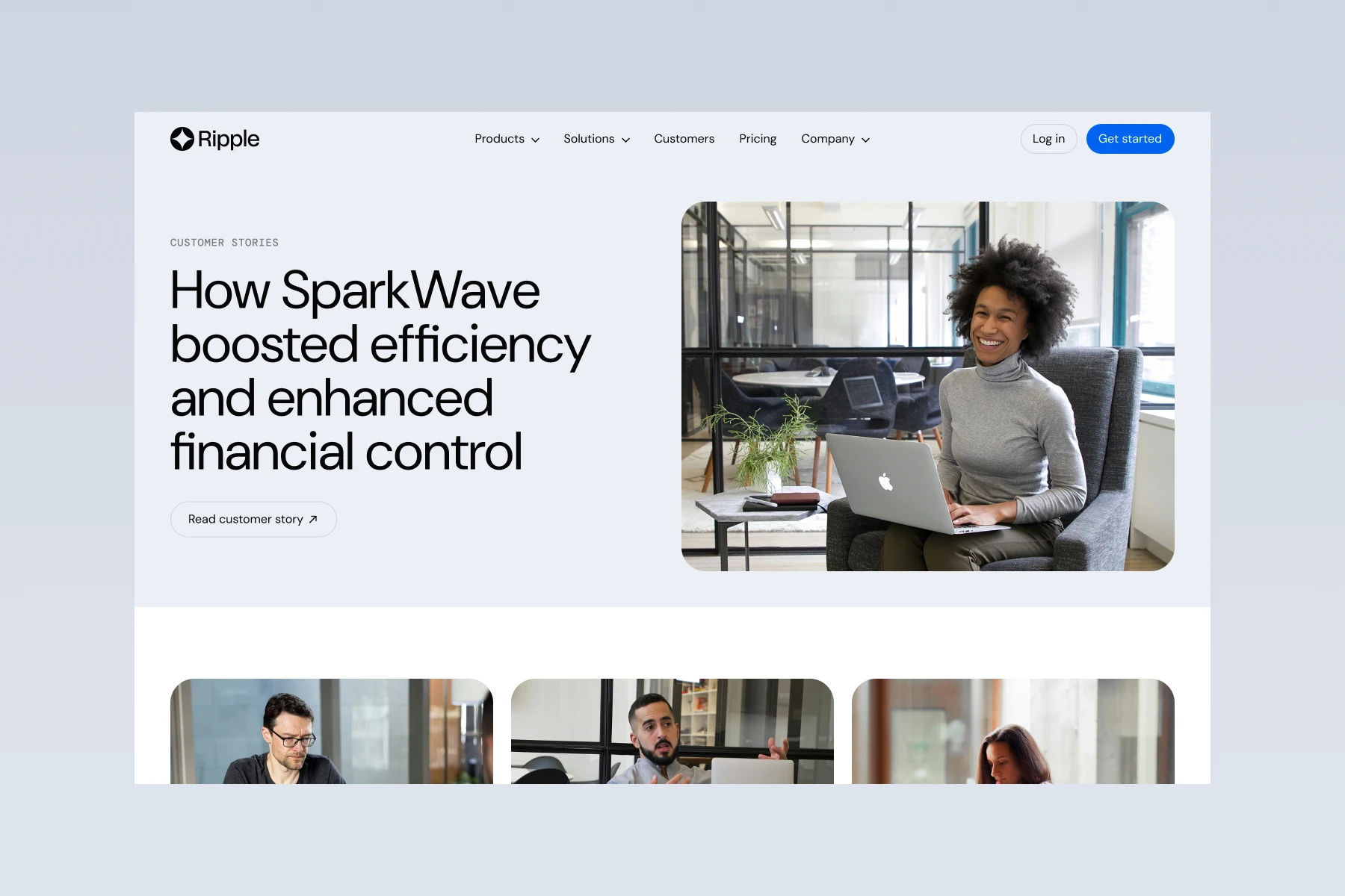 A smiling woman with curly hair sitting in an office chair working on a laptop in a modern office with glass partitions.
