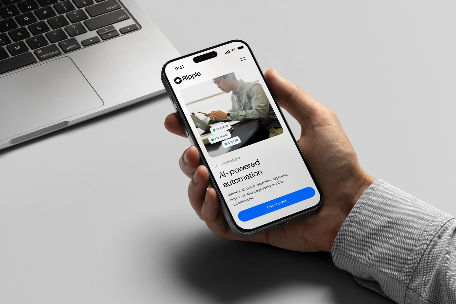 Hand holding smartphone displaying Ripple app with AI-powered automation and a Get started button, next to a laptop keyboard.