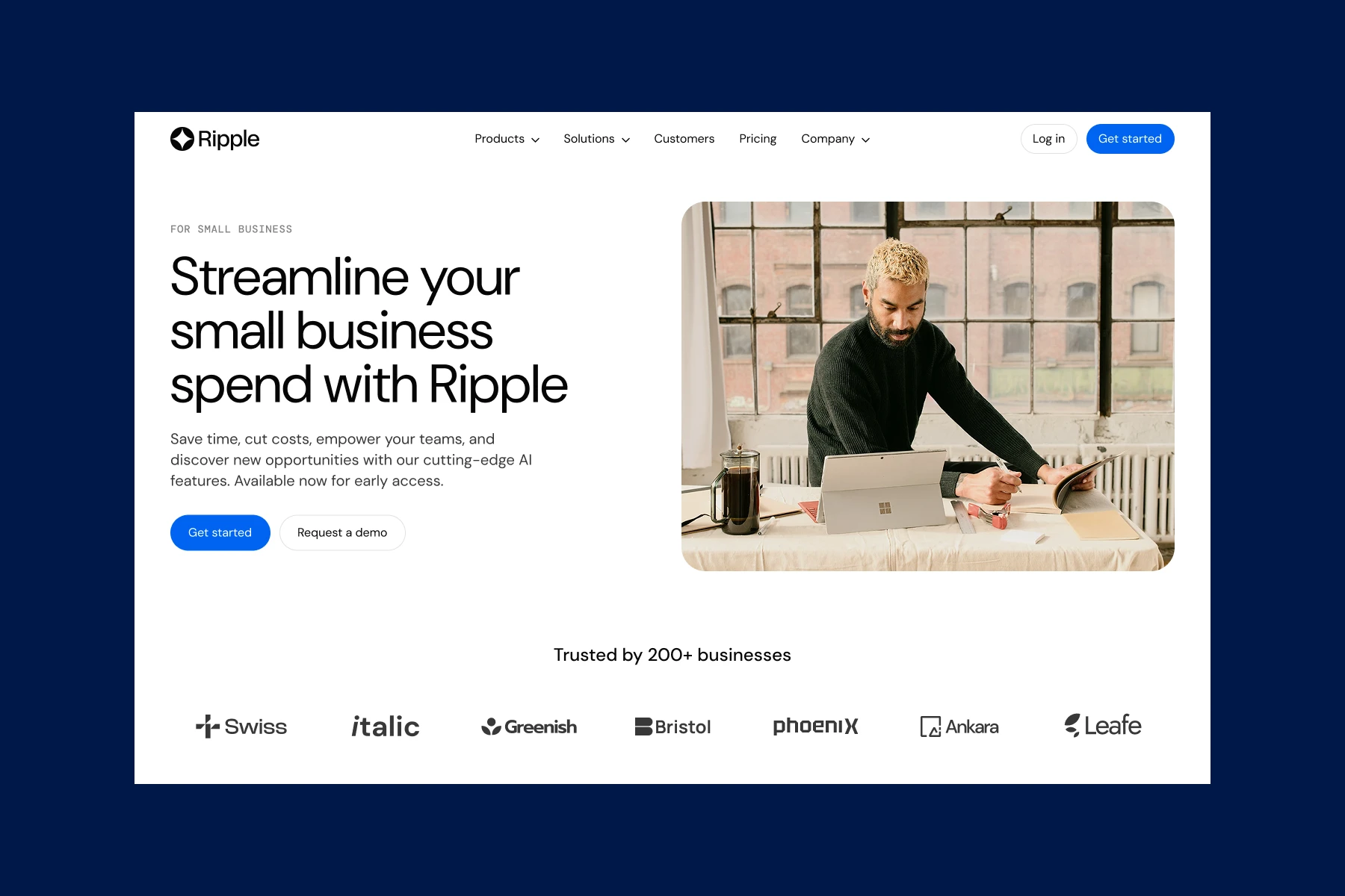 Man with blond hair and beard working on tablet and writing in notebook at a table near large windows.