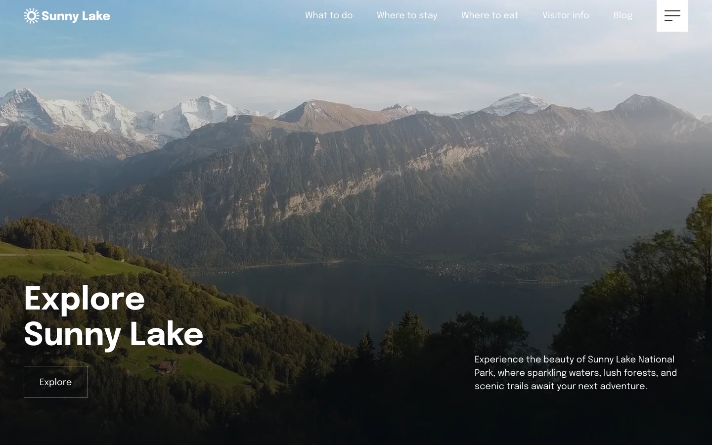 View of a large lake surrounded by green hills and tall mountains with snow-capped peaks under a clear sky.