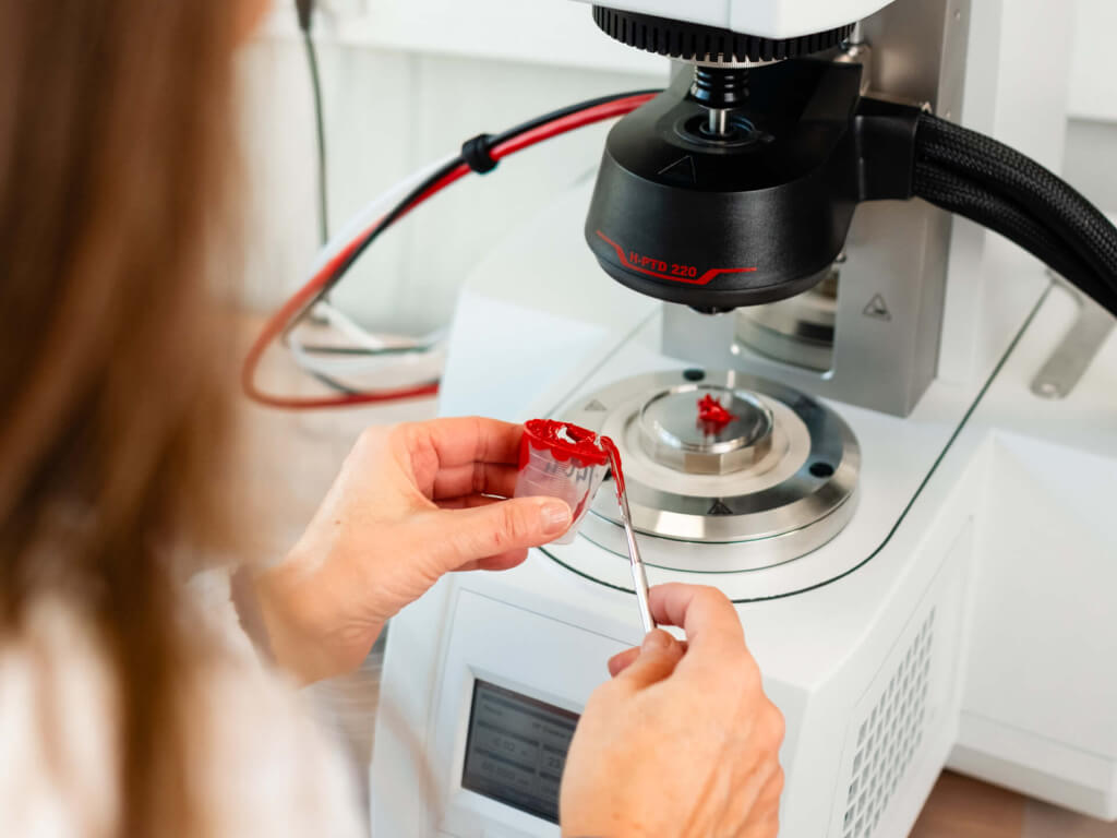 Person hält einen Behälter mit rotem Material und verwendet ein Werkzeug neben einem Analysegerät in einem Labor.