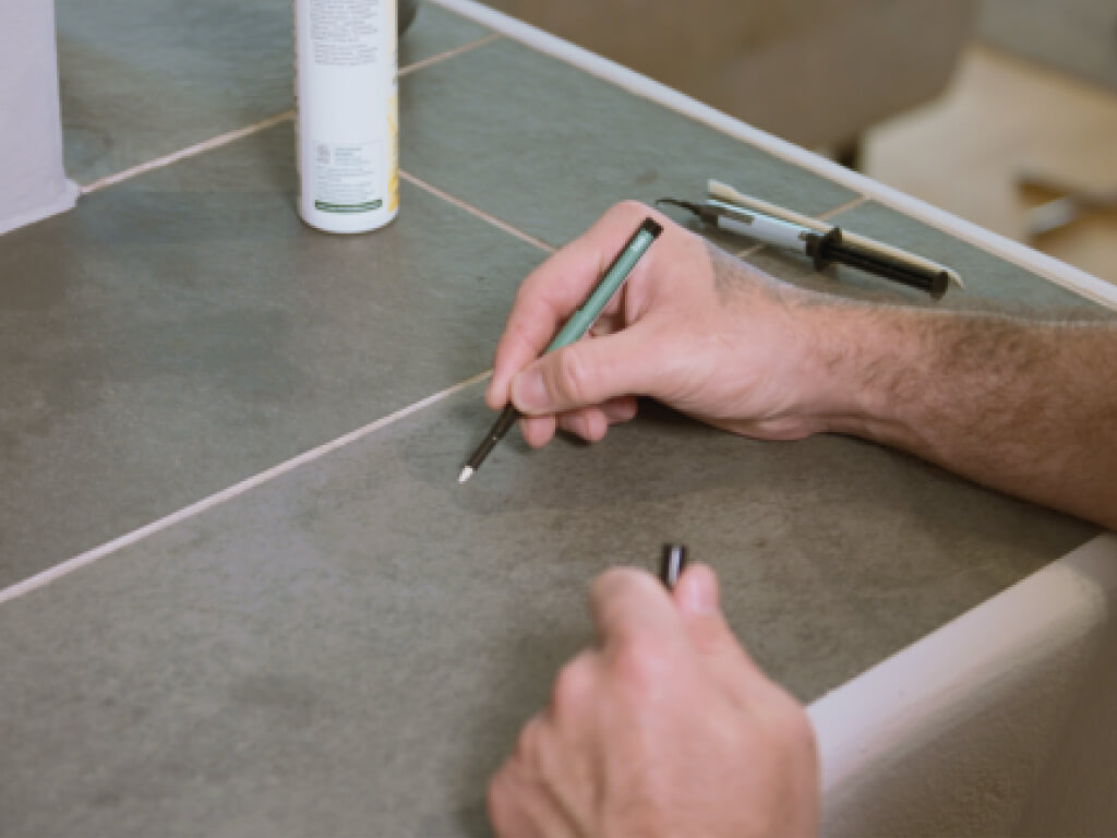 Eine Hand benutzt einen stonelux reparatur Stift, um Fugen zwischen grauen Fliesen auf einer Arbeitsfläche auszubessern.