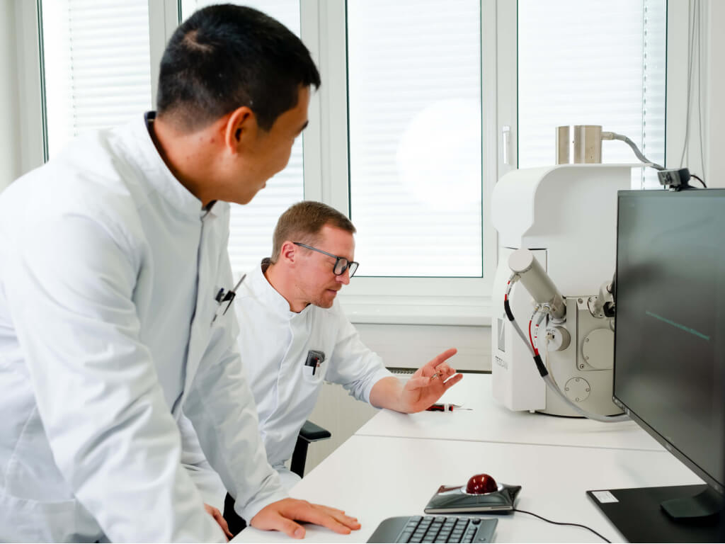 Zwei Wissenschaftler in weißen Kitteln bedienen ein Elektronenmikroskop und betrachten den Monitor in einem Labor.
