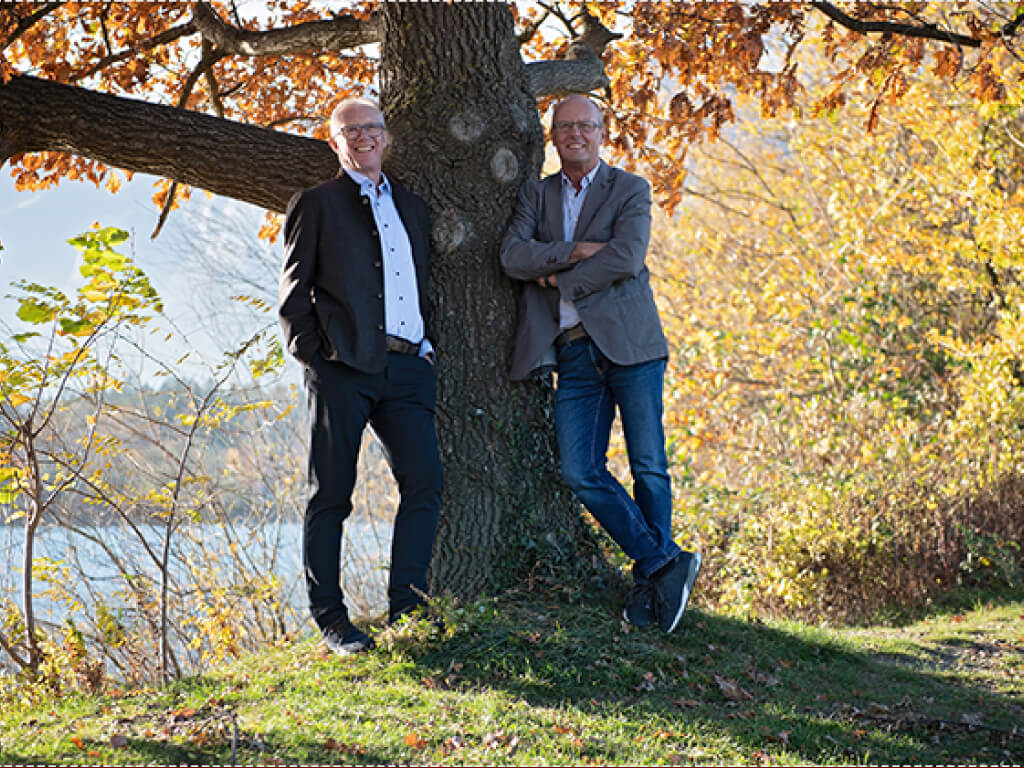 Zwei Männer lehnen an einem Baum mit herbstlich gefärbten Blättern in einem Park.