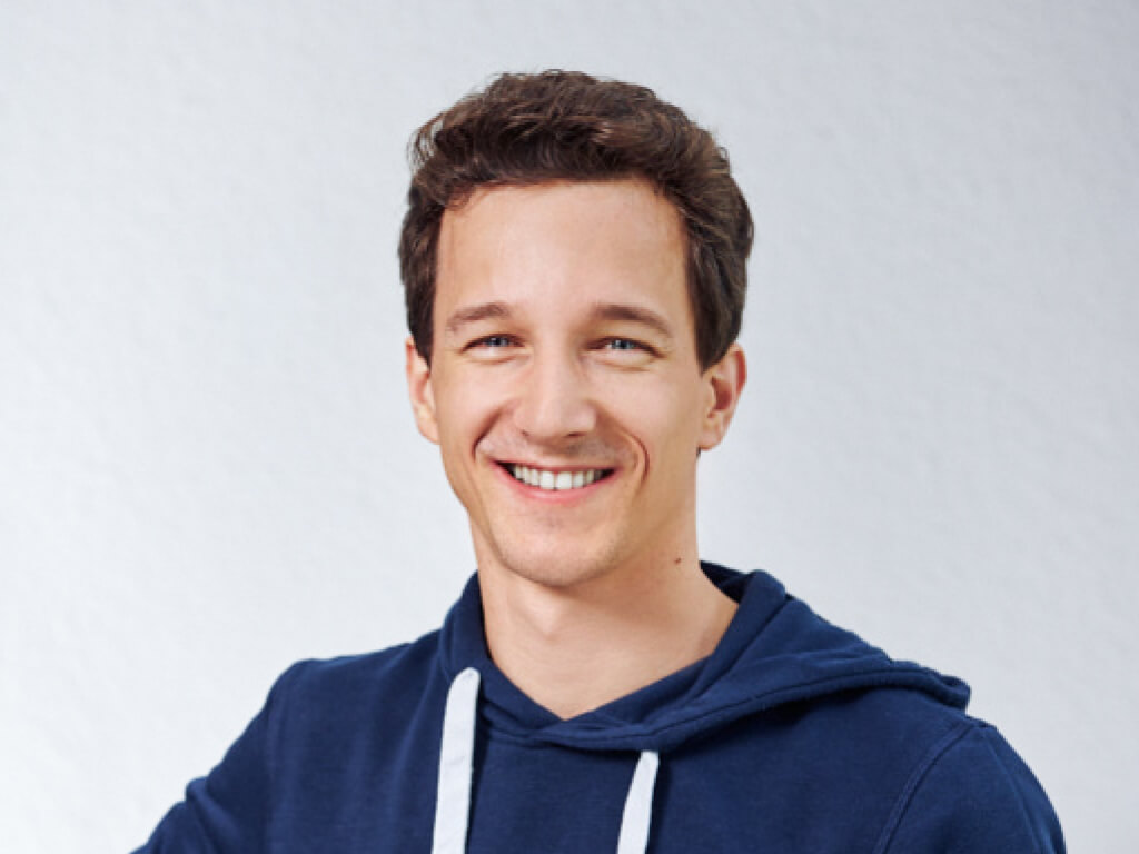 Mann mit kurzen braunen Haaren, der in einem dunkelblauen Kapuzenpullover lächelt.