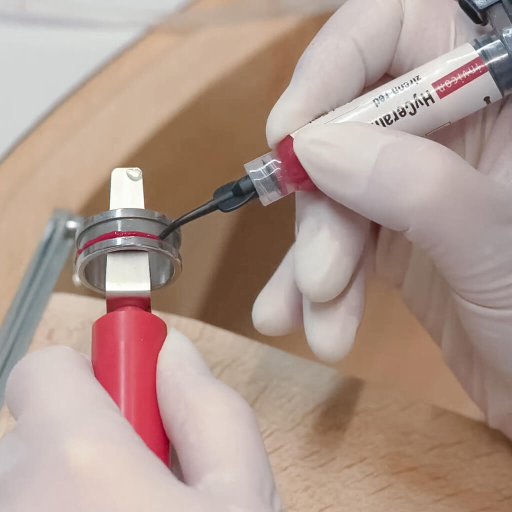 Eine Person mit weißen Handschuhen trägt rotes hyceram mit auf ein Metallring-Werkzeug mit rotem Griff auf.