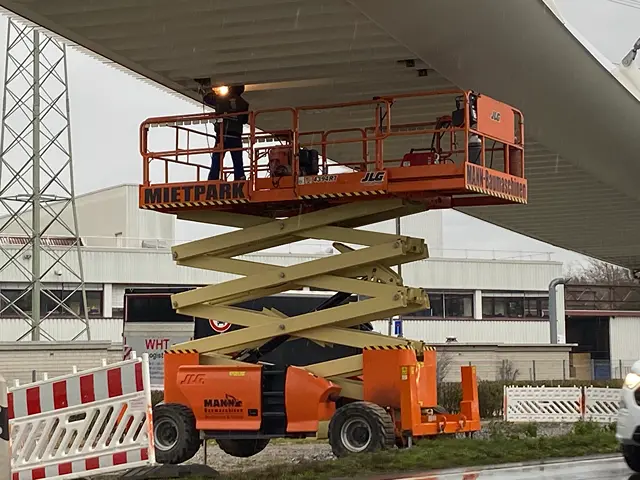 Eine orangefarbene Scherenarbeitsbühne mit der Aufschrift 'MIETPARK' und 'MANN Baumaschinen', auf der eine Person arbeitet unter einem großen Dach.
