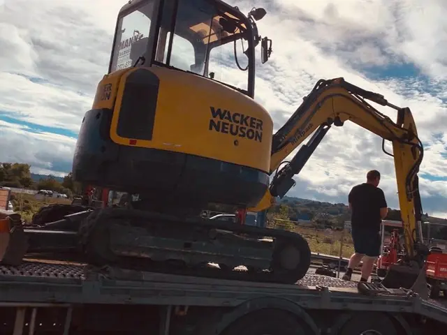 Gelber Wacker Neuson Minibagger auf einem Tieflader mit einem Mann daneben bei bewölktem Himmel.