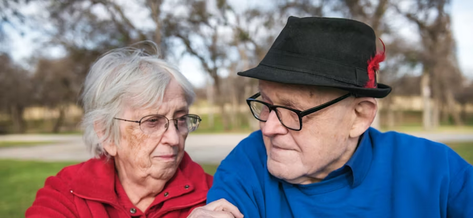 Elderly woman in red jacket looking affectionately at elderly man in blue sweater and black hat with red feather outdoors.