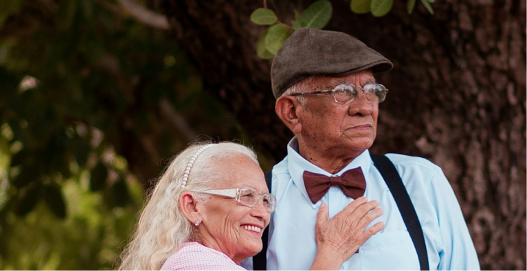 Elderly couple outdoors, woman with white hair smiling and resting her hand on man's chest, man wearing a cap, bow tie, glasses, and suspenders.
