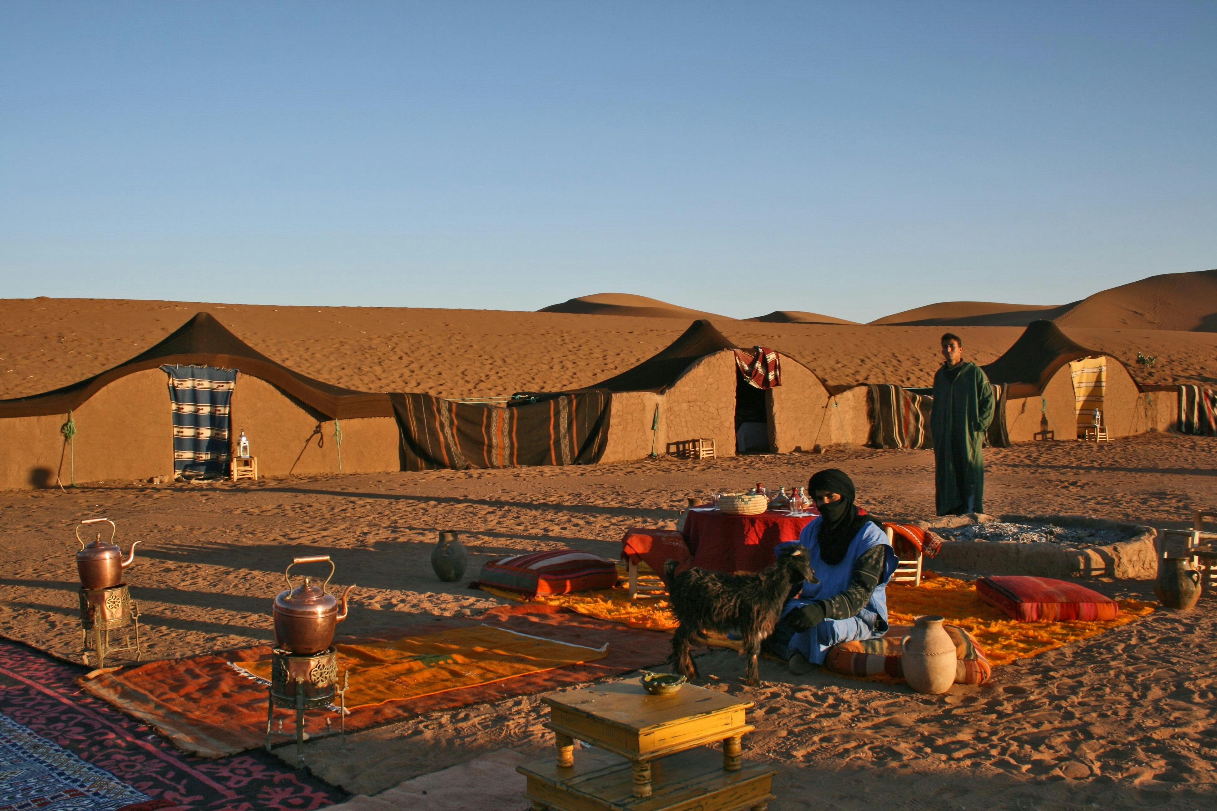 Two men in traditional robes and headwraps sitting and standing around a camp setup with rugs, cushions, clay pots, and kettles in a desert with mud tents and sand dunes in the background.