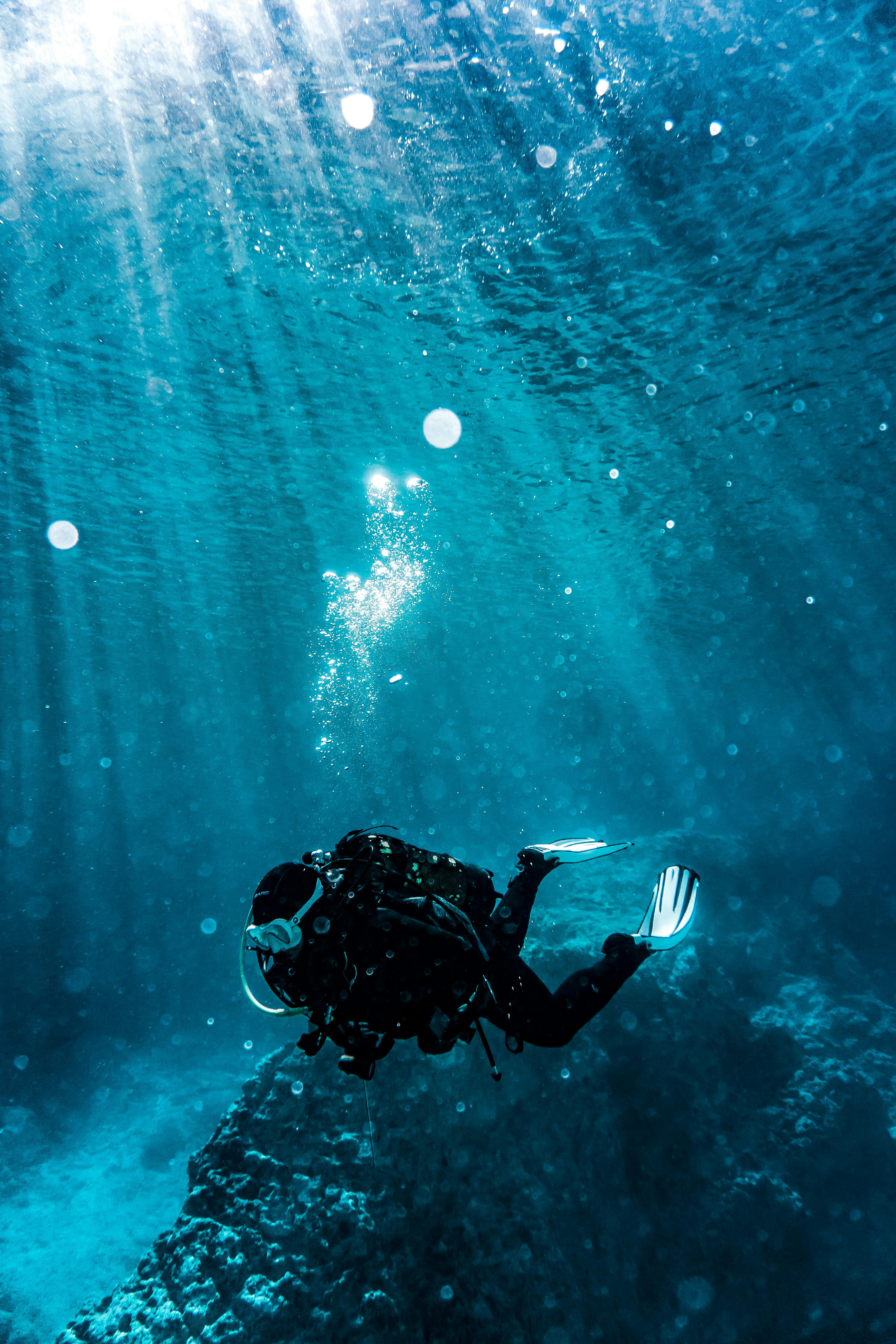 Scuba diver underwater surrounded by blue water and bubbles near a rocky seabed.