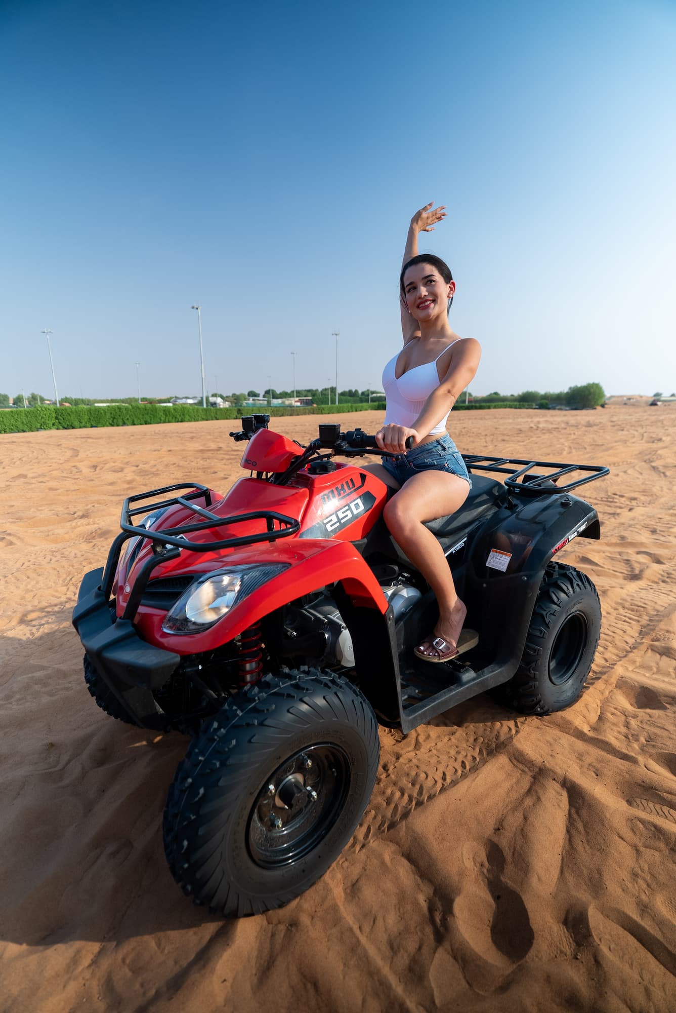 Smiling woman in white tank top and denim shorts sitting on a red ATV in a sandy desert area under a clear blue sky.