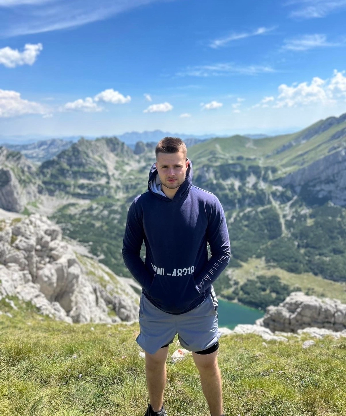 Man in navy hoodie and gray shorts standing on grassy hill with rocky mountains and lake in the background.