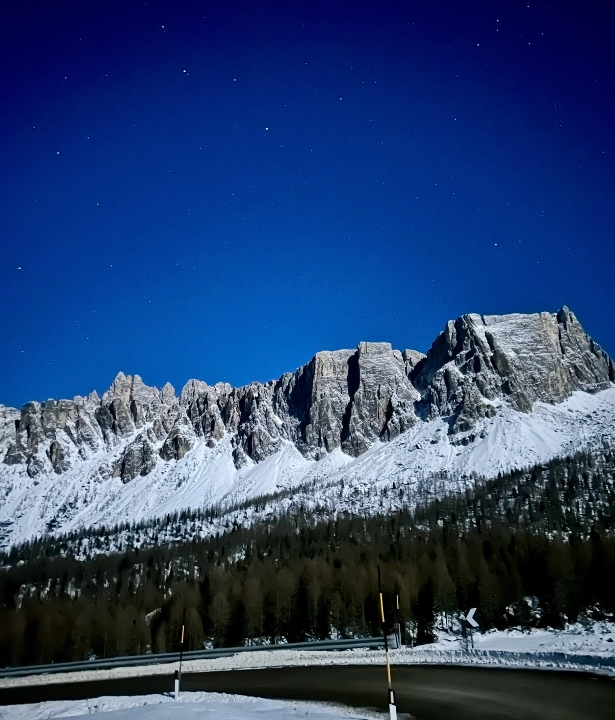 Snow-covered mountain range under a dark blue night sky dotted with stars, with a snowy road in foreground.